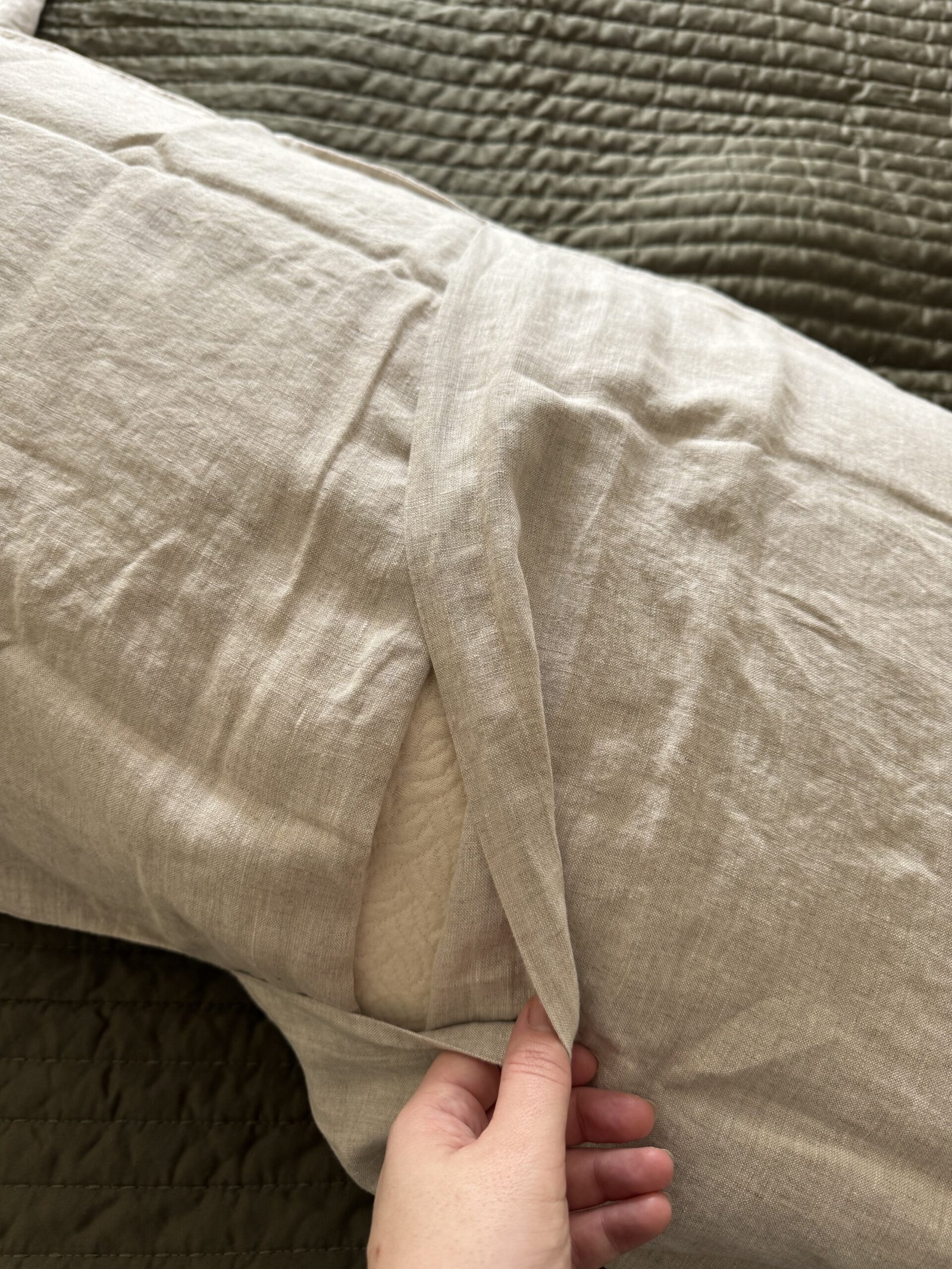 Hand adjusting the corner of a light beige pillowcase on a pillow, showing the interior fabric. The pillow is on a textured green surface.