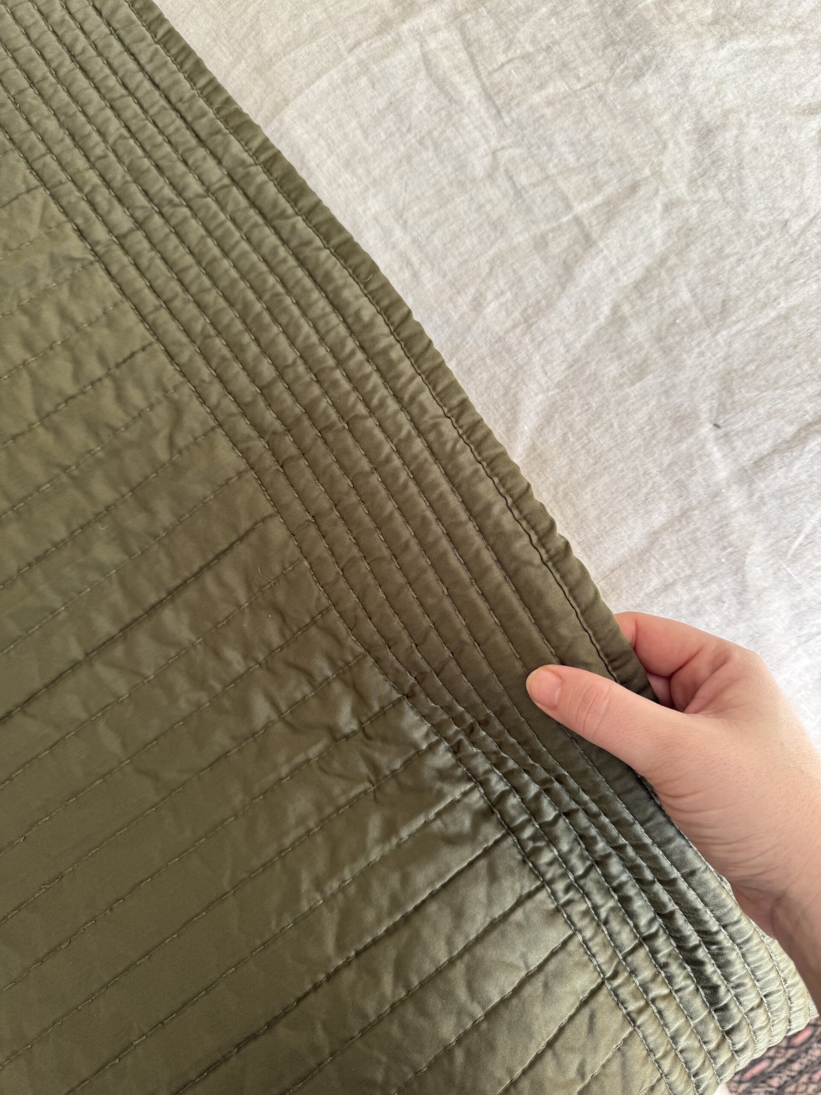 A hand holds the corner of a quilted fabric with parallel stitching. The fabric is a smooth, olive green material against a light-colored textured background.