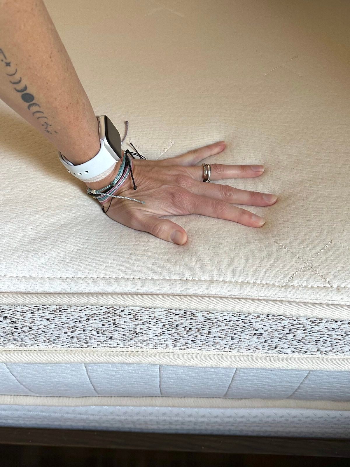 Reviewer Randi Donahue's hand pressing down on an organic Birch mattress, demonstrating its texture and responsiveness. The person is wearing a smartwatch and several bracelets, making it clear this isn't just a mattress review but a glimpse into lifestyle choices aligned with comfort and sustainability.