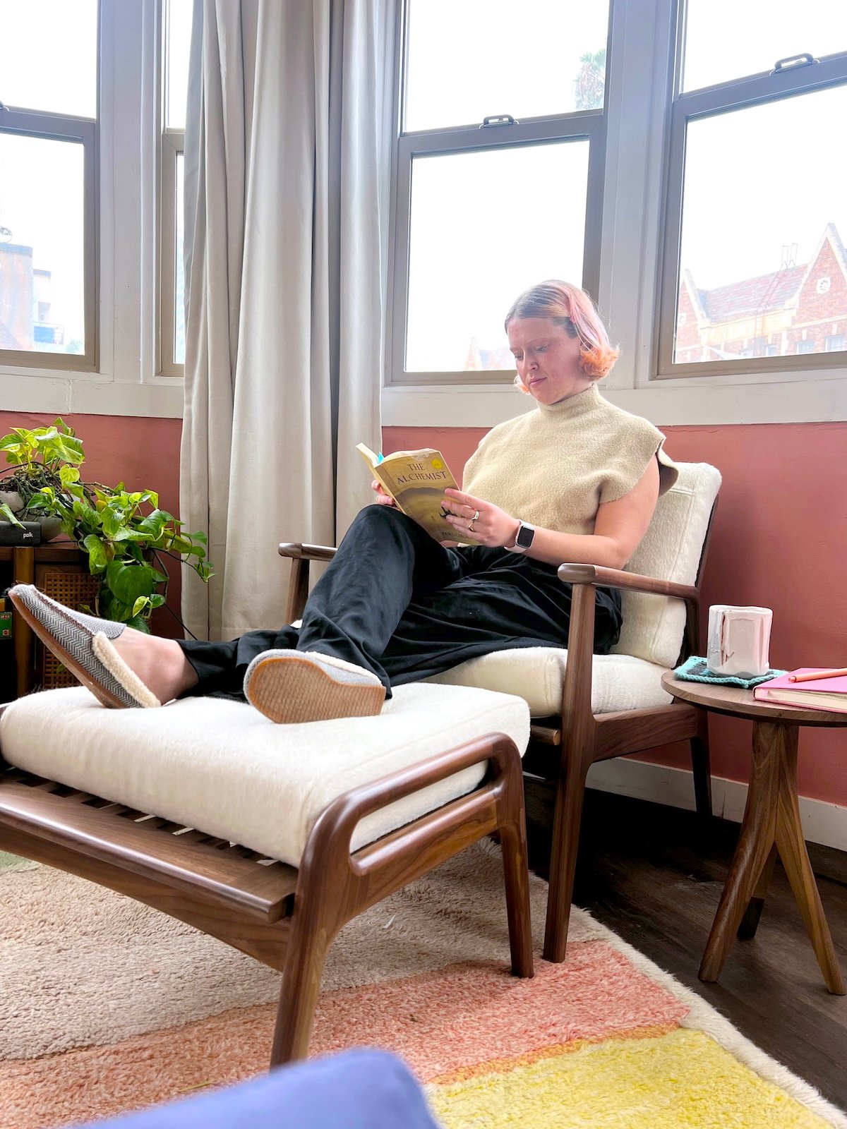 Person with short hair sitting on a wooden lounge chair, reading a book. There are plants nearby and a mug on a side table. Sunlight streams through the windows.