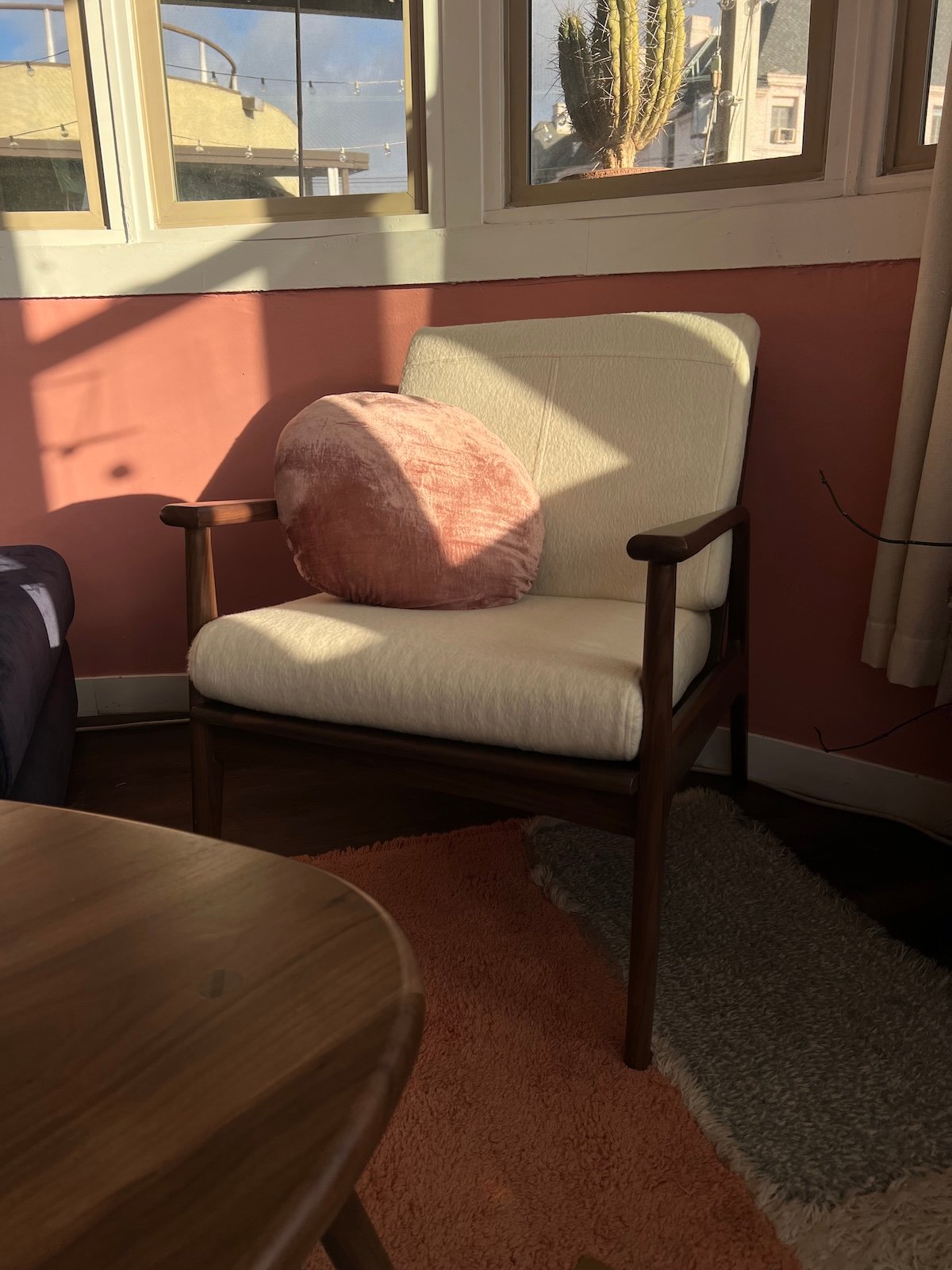 A white cushioned chair with a round pink pillow is placed next to a window in a sunlit room with pink walls.