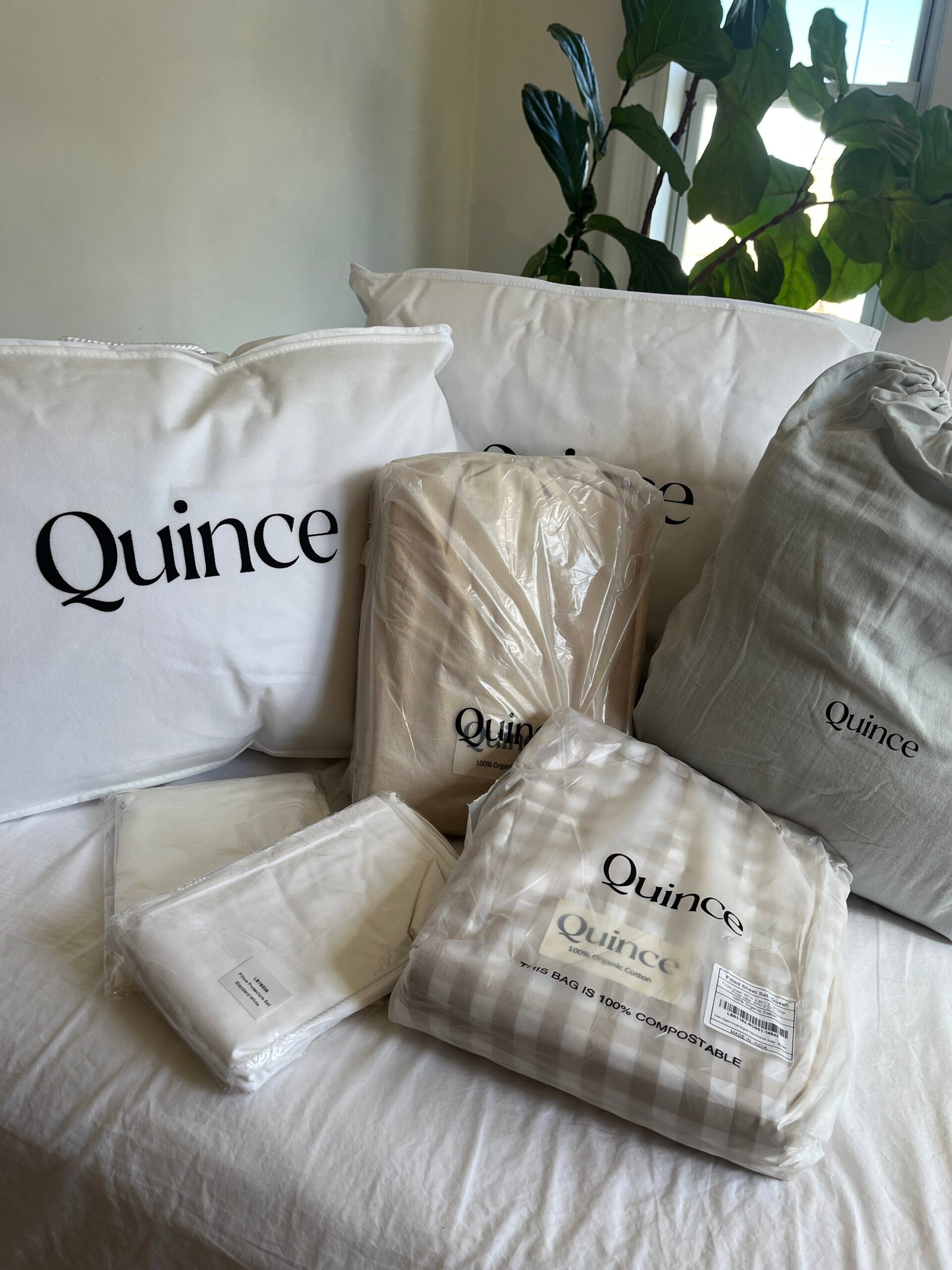 Quince branded bedding items, including pillows, blankets, and sheet sets, arranged on a bed. A leafy plant is visible in the background.
