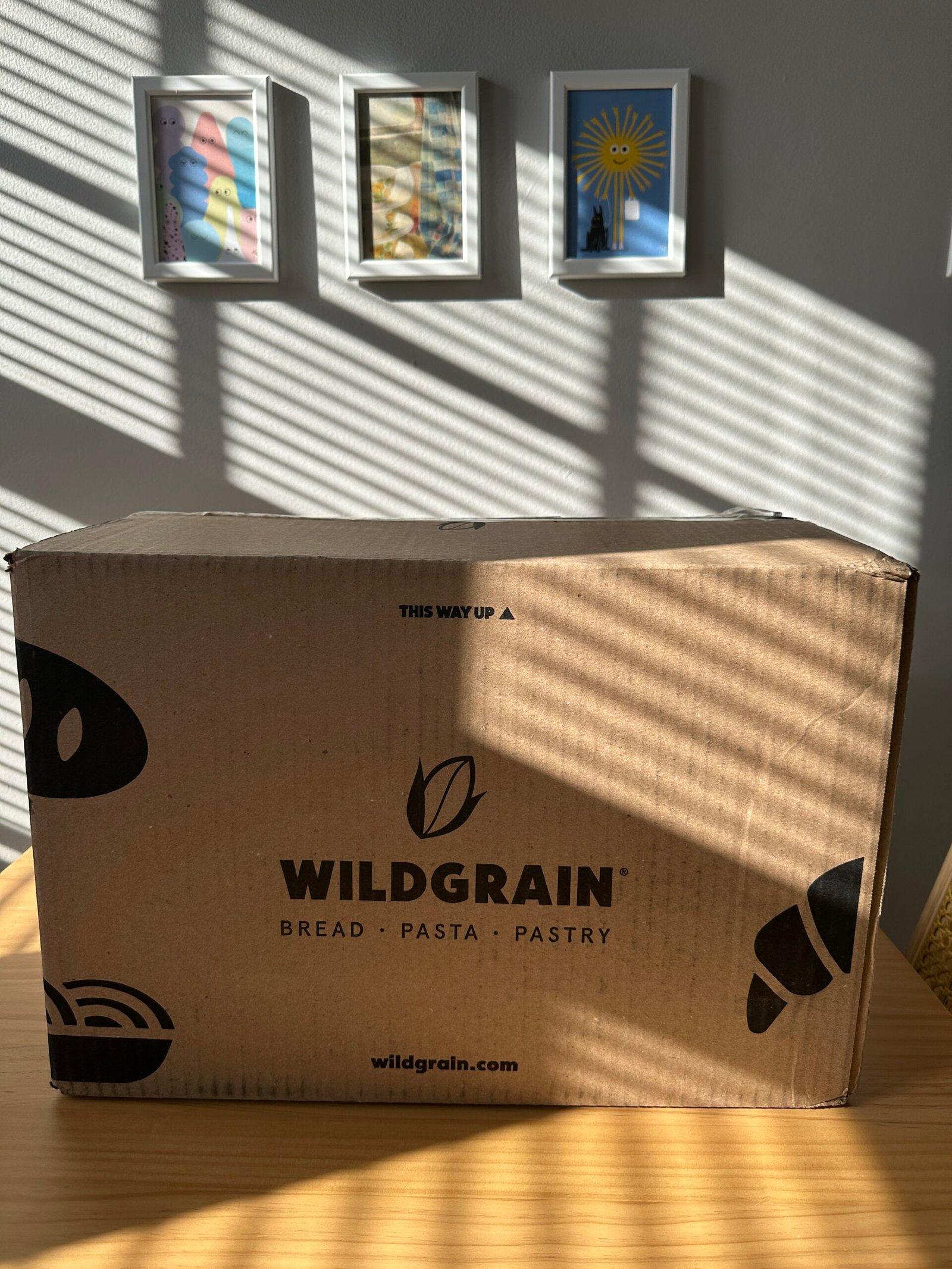 A large cardboard box labeled "Wildgrain Bread Pasta Pastry" sits on a sunlit table, with three framed pictures on the wall behind it.