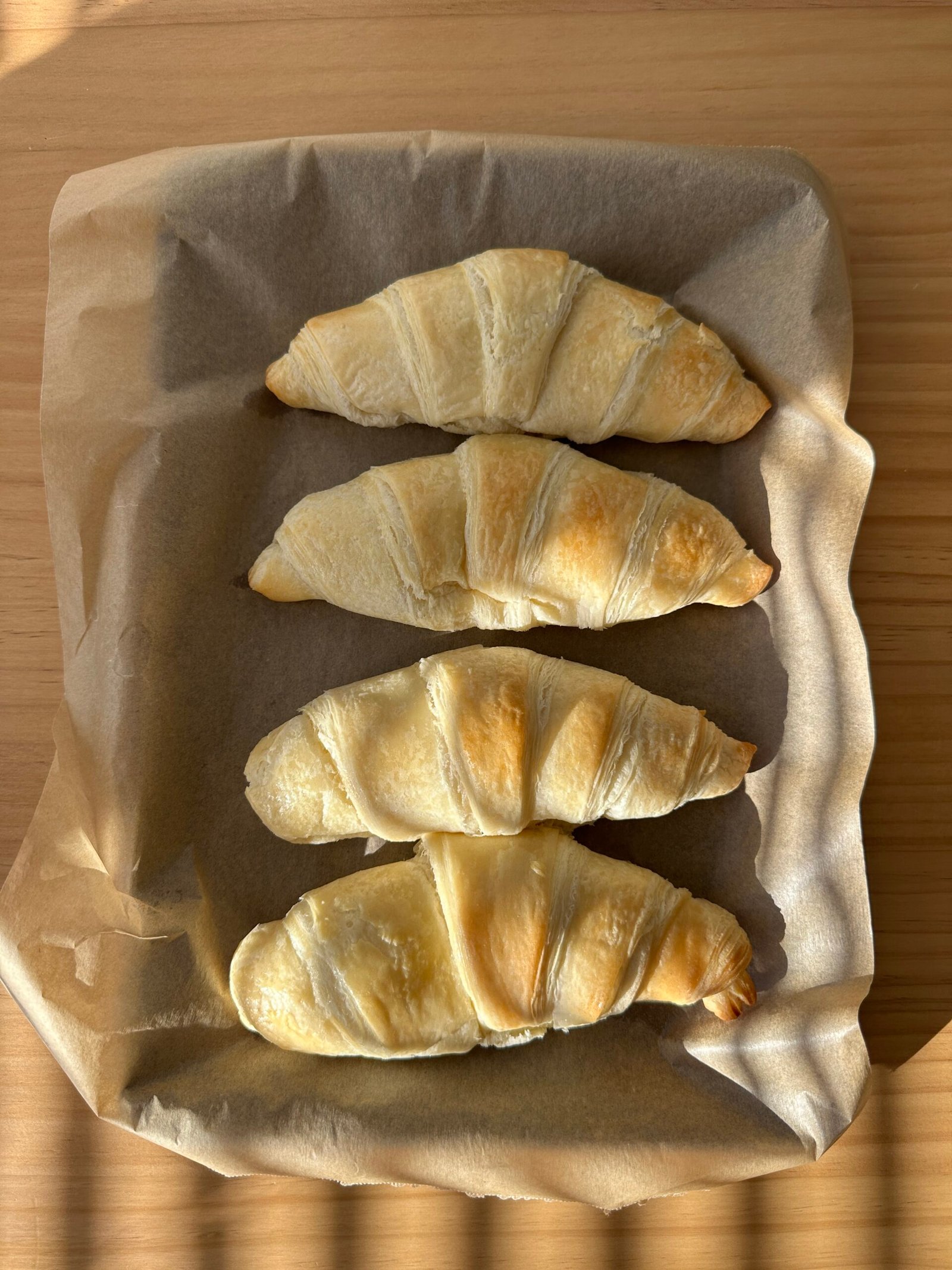 Four golden brown croissants on a baking sheet lined with parchment paper, placed on a wooden surface.