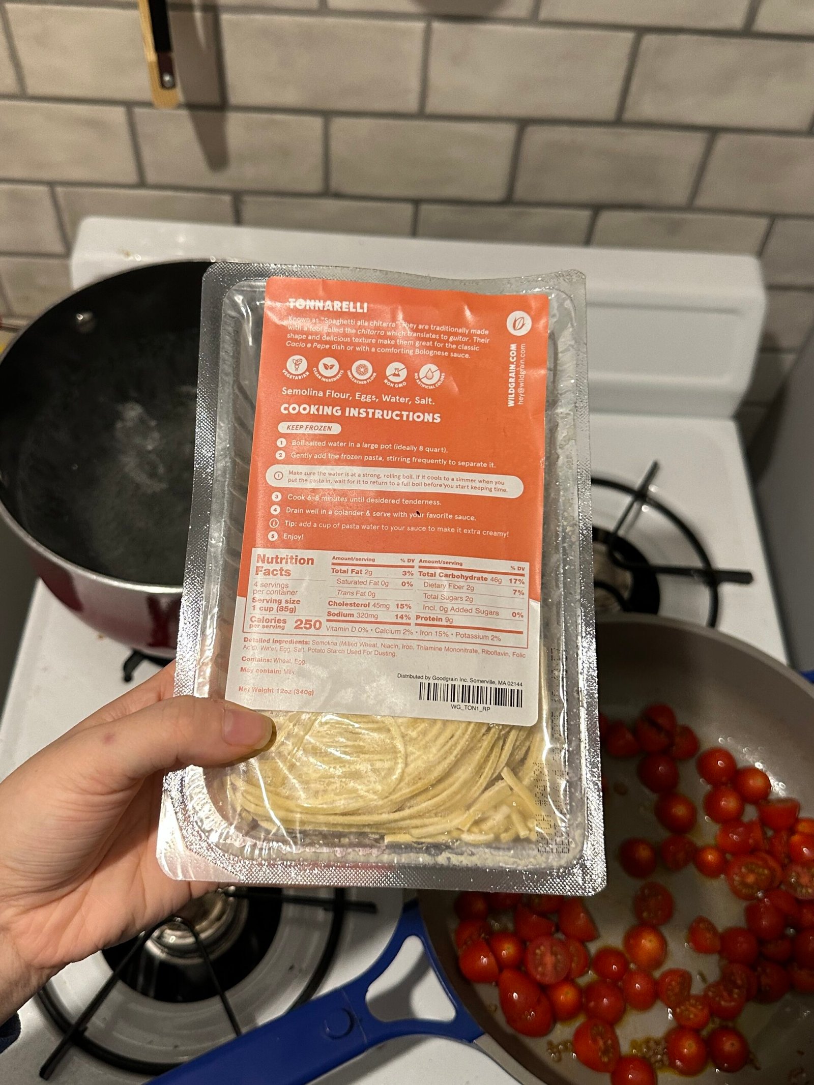 A hand holds a package of spaghetti noodles in a kitchen. Nearby is a pot of boiling water and a pan with cherry tomatoes.