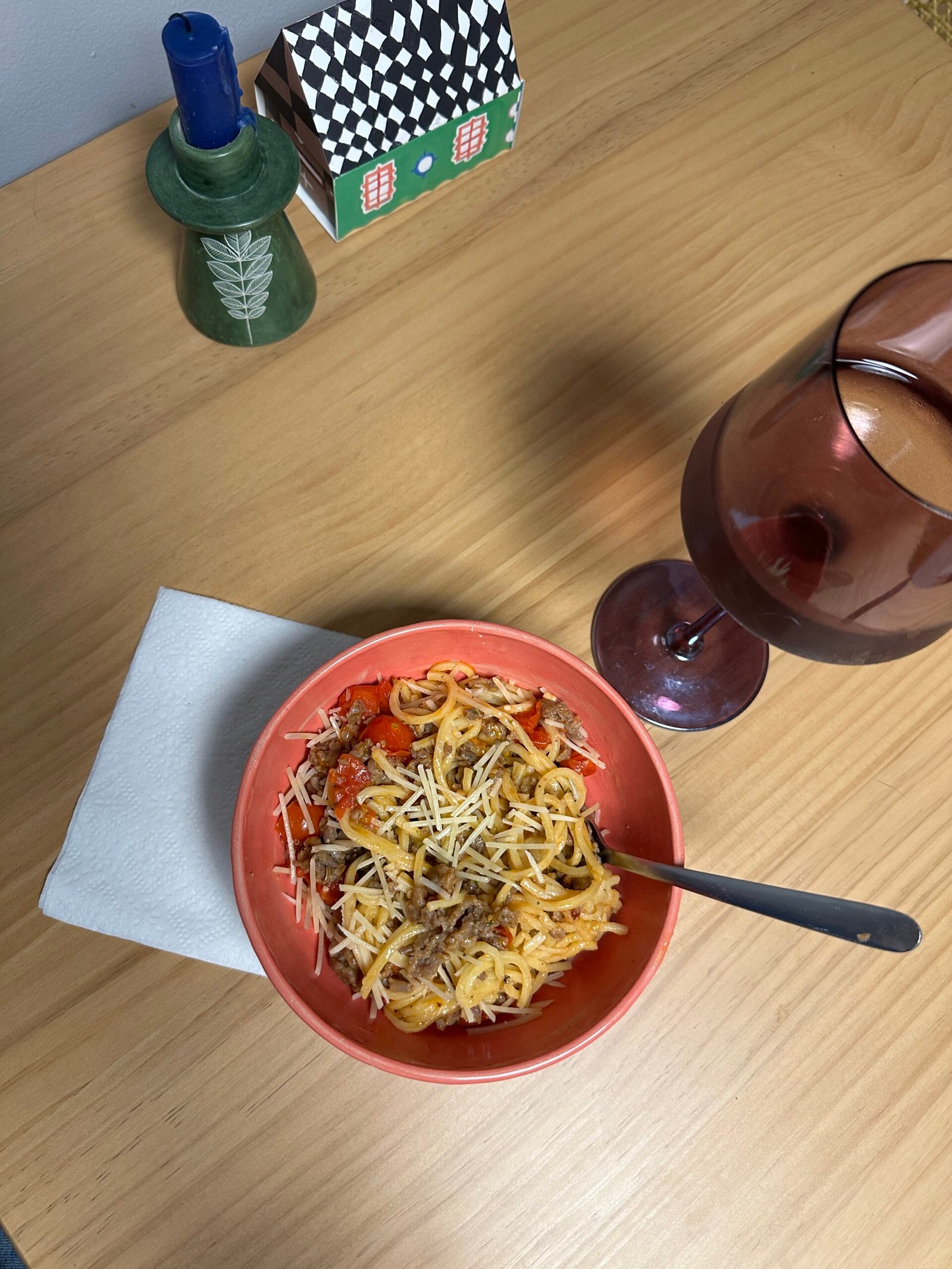 Bowl of spaghetti with meat sauce and cheese on a table, beside a glass of red wine, a candle, a napkin, and a small decorative house.