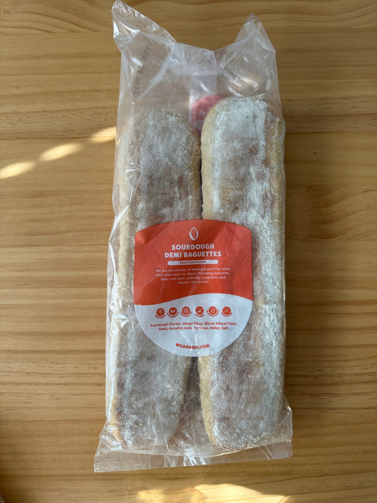 Two packaged sourdough demi baguettes in a clear bag with an orange and white label on a wooden surface.