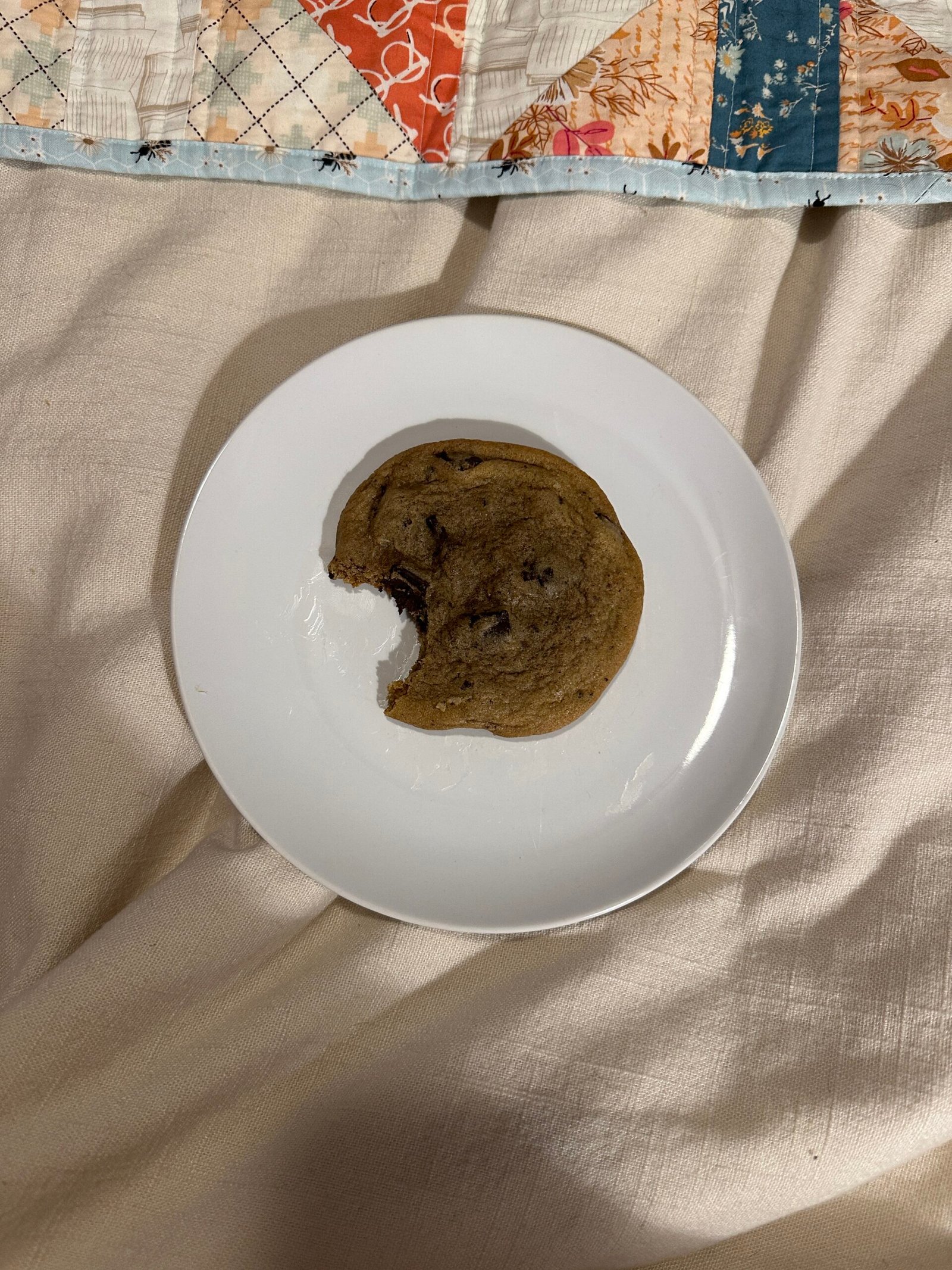 A chocolate chip cookie with a bite taken out of it sits on a white plate on a quilted fabric surface.