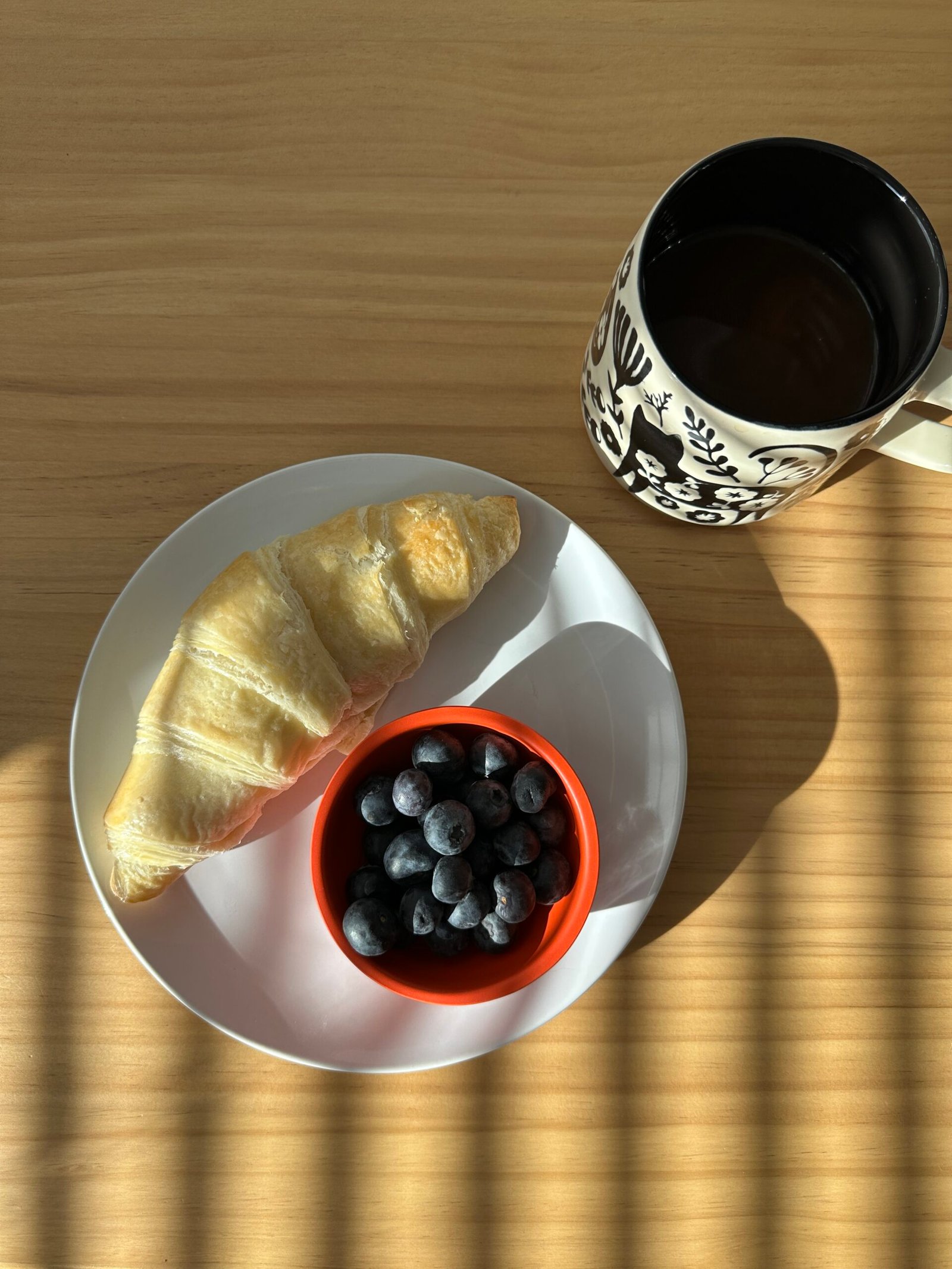 A croissant and a small bowl of blueberries on a white plate next to a patterned mug of coffee on a wooden table. Sunlight casts shadow lines across the scene.