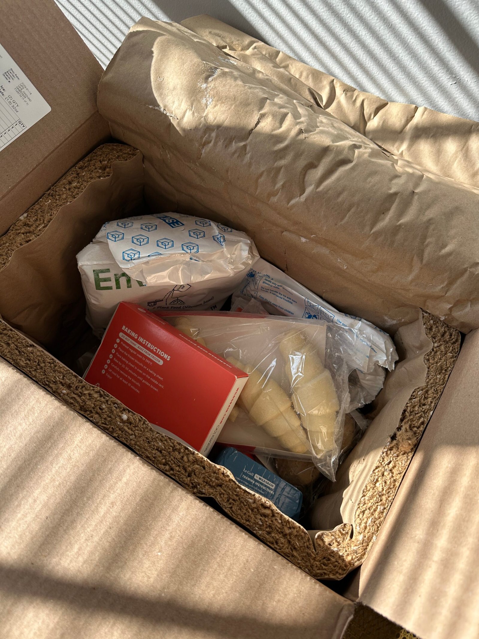 Open cardboard box containing insulated packaging and various food items, including sealed bags and a red booklet, with sunlight casting shadows.