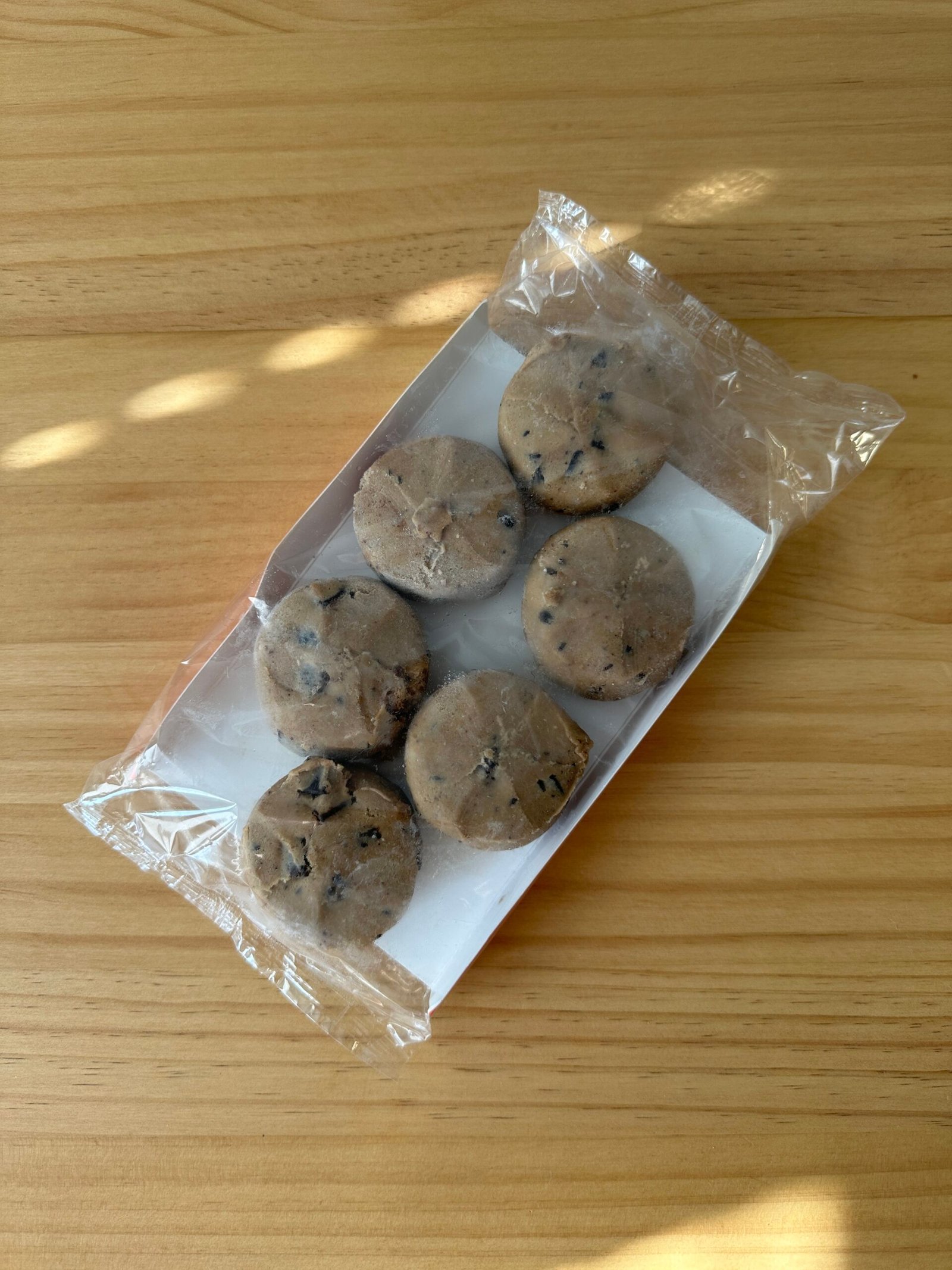 Six round cookies with chocolate chips in an open plastic tray, placed on a wooden surface.