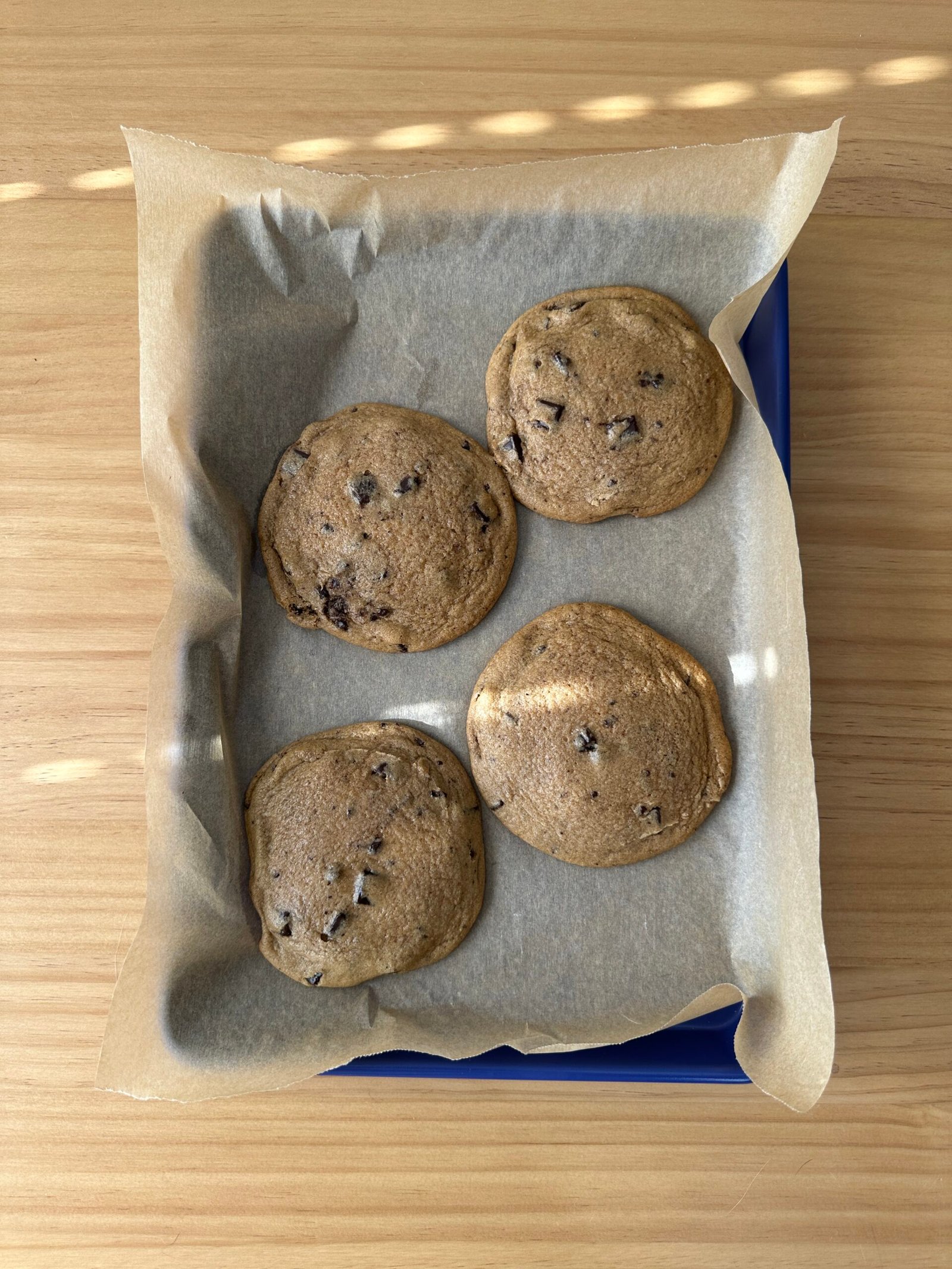 Four chocolate chip cookies on parchment paper in a baking tray on a wooden surface.