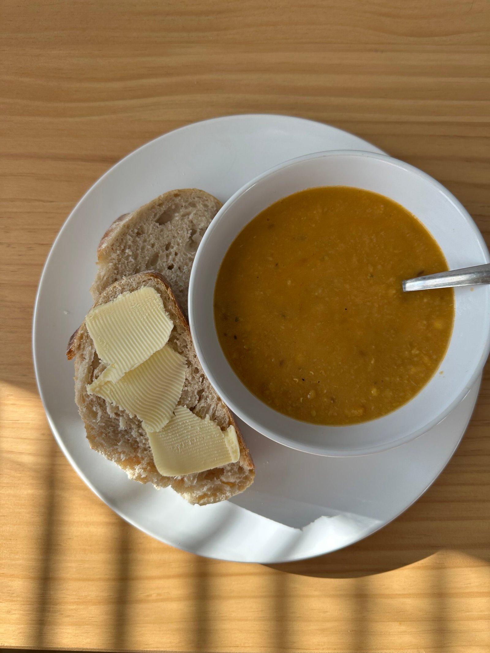 Bowl of orange soup with a spoon, served on a white plate with a slice of bread topped with butter on a wooden table.