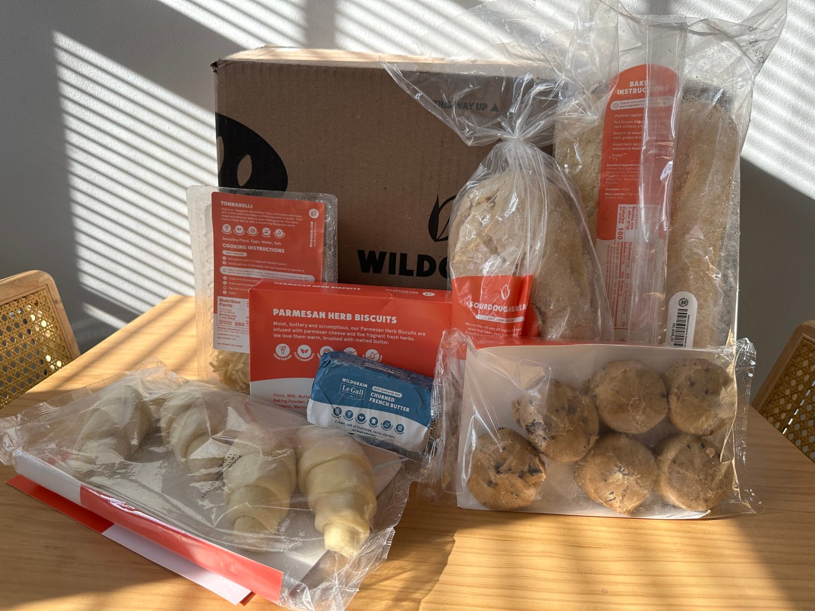 A Wildgrain subscription box with various packaged baked goods including croissants, bread, and biscuits displayed on a wooden table.