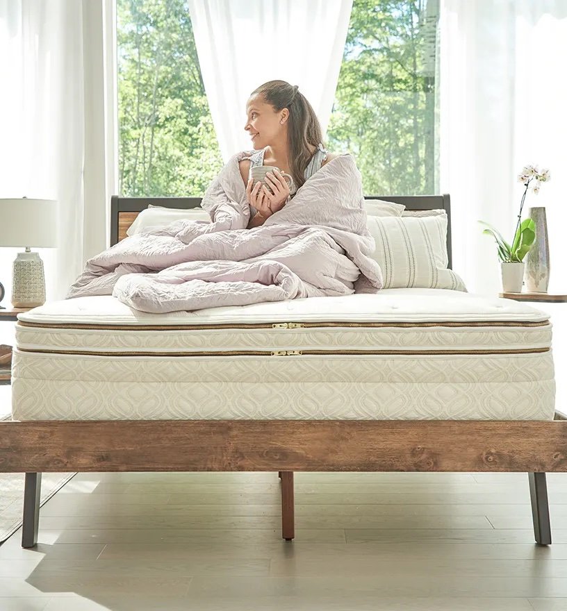 A person sits on a Naturepedic mattress with a light comforter, holding a mug, in a sunlit room. Large windows offer a view of trees outside.