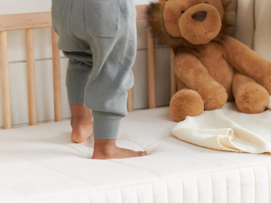 A toddler wearing gray pants stands on an organic mattress next to a plush lion toy.