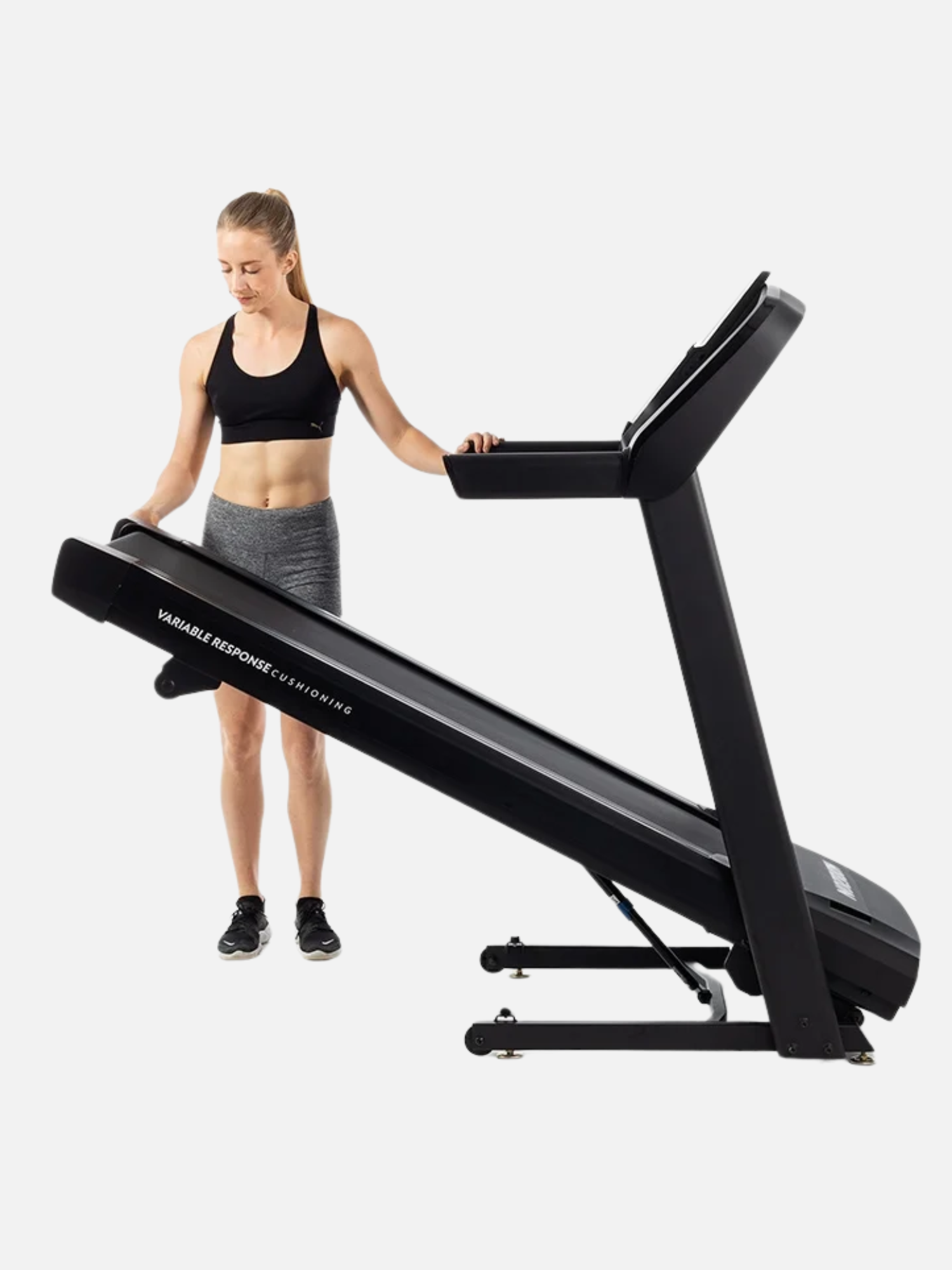 A woman in workout attire is adjusting a treadmill to fold for storage.