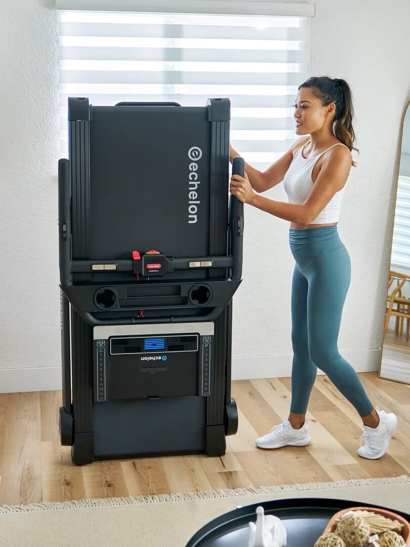 Person setting up a folded treadmill in a living room with a mirror, couch, and window blinds visible.