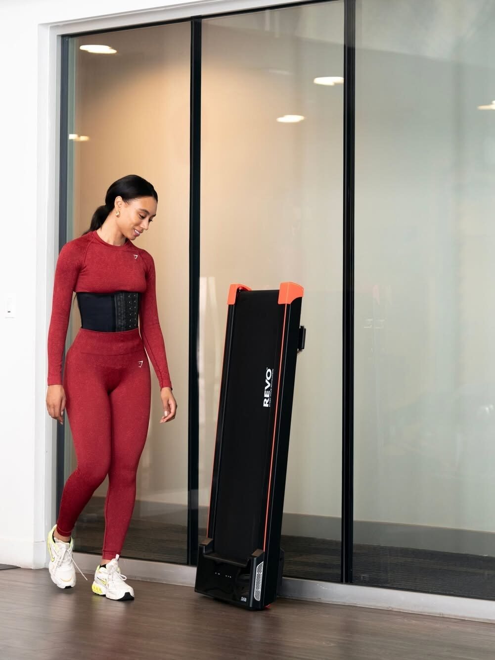 A person in a red athletic outfit walks past a folded treadmill in front of glass doors.