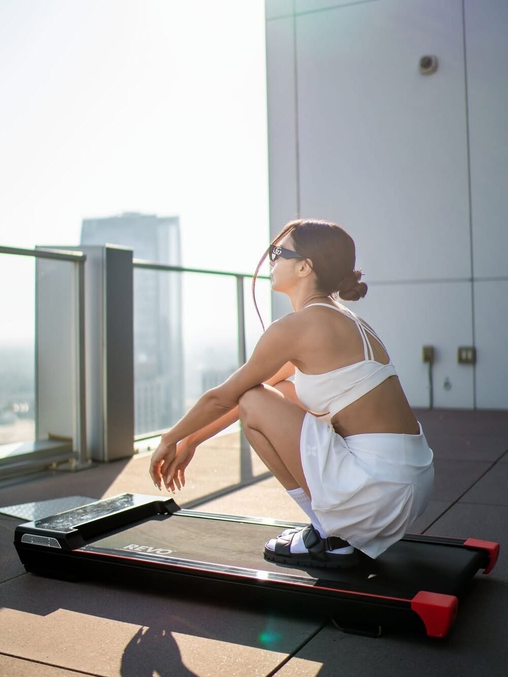 Person squatting on a treadmill on a rooftop, wearing a white outfit and sunglasses, with a cityscape in the background.