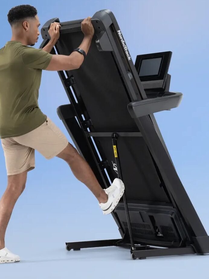 A man in a green shirt and shorts folds a treadmill deck upwards. Text above reads, "Maximize your room’s space with an easy-to-fold deck.