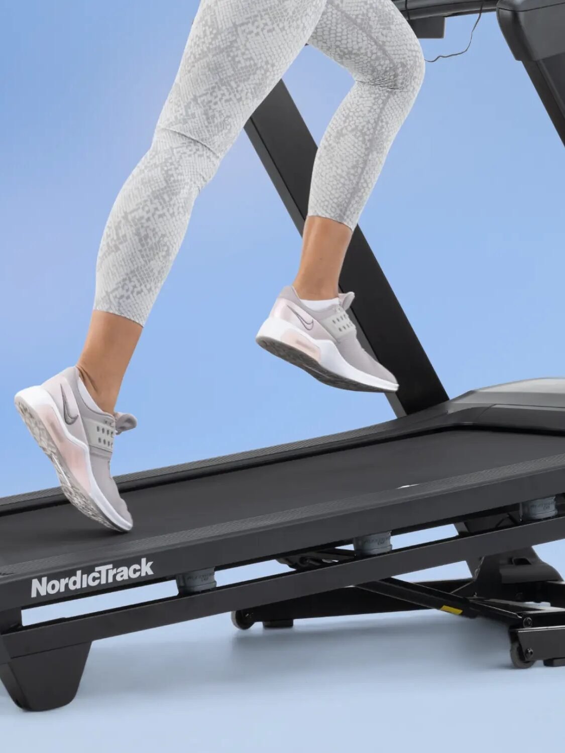 Person running on a NordicTrack treadmill, wearing light grey patterned leggings and white sneakers, against a light blue background.