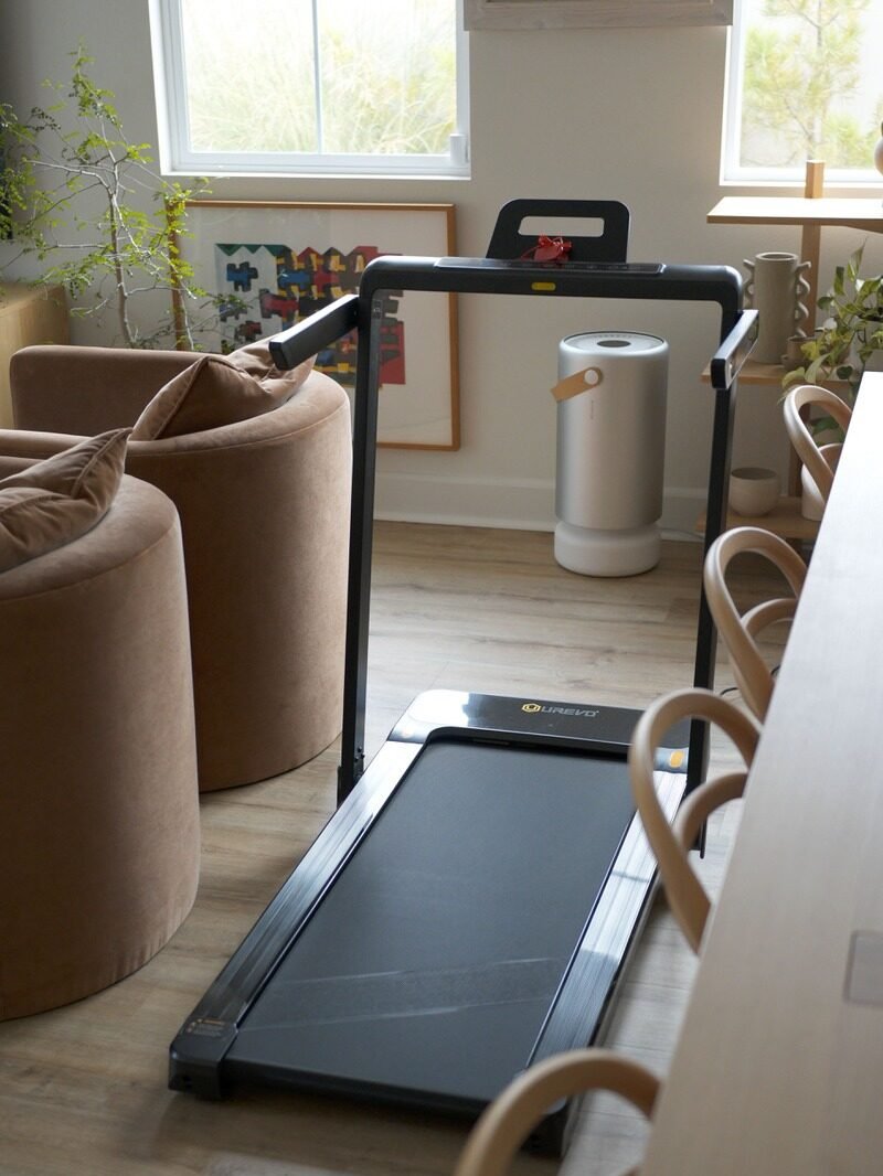 A treadmill is placed indoors near brown chairs and light wooden furniture, with a modern air purifier in the background.