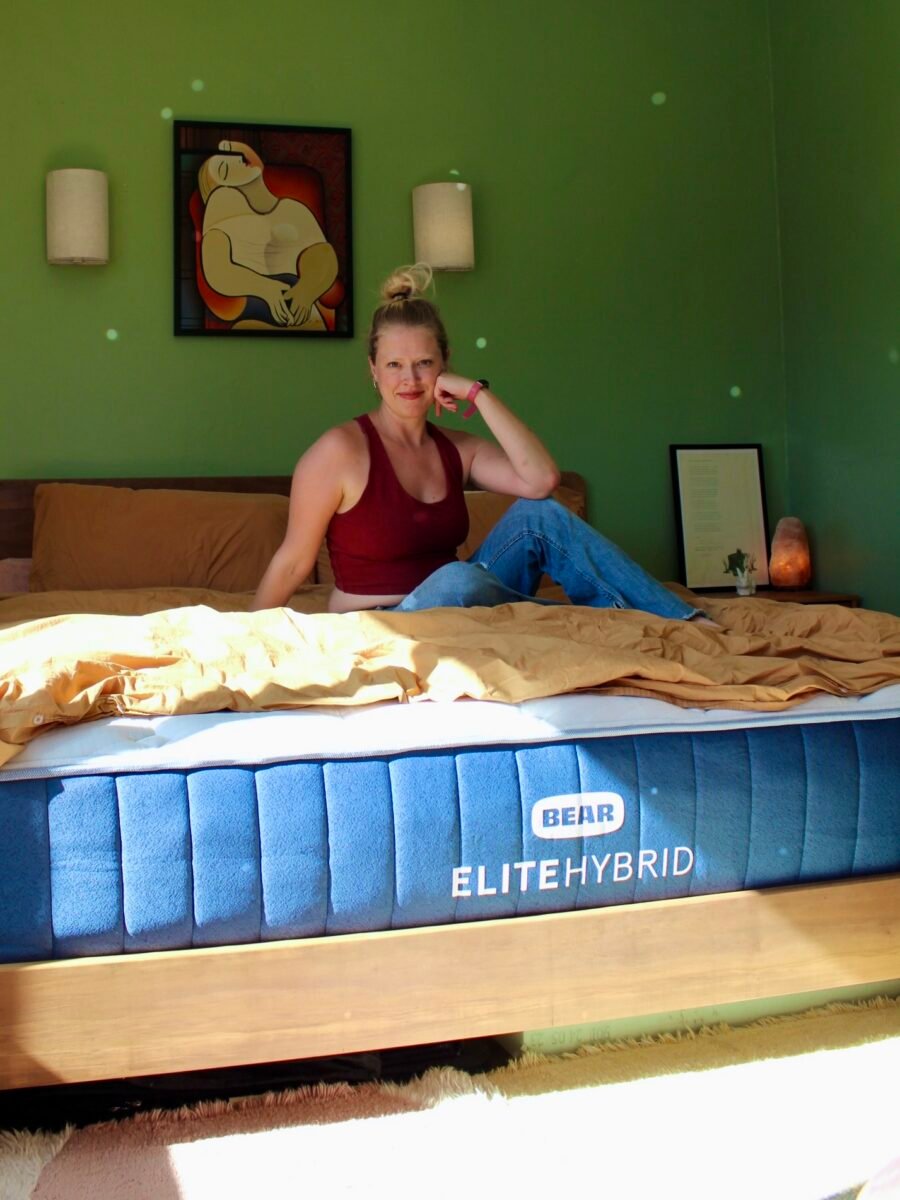 A person sitting on a bed with a "Bear Elite Hybrid" mattress in a green room, wearing a red top and jeans. A picture is on the wall above, and a lamp is on a side table.