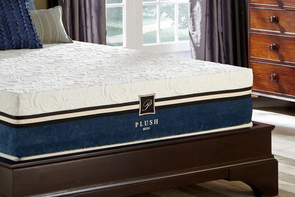 A bedroom scene featuring a plush mattress with a decorative white swirl pattern, resting on a dark wood bed frame. Partially visible in the background are a dresser and large windows.