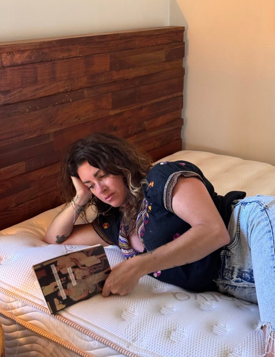 Person lying on a bed, propped on one elbow, reading a magazine. They are wearing a black shirt and jeans, in a room with a wooden headboard.