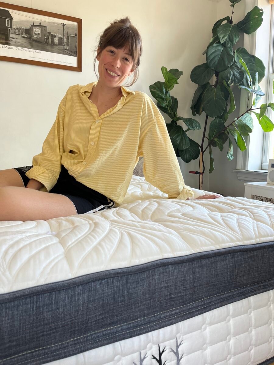 Person in a yellow shirt sitting on a bed with a white and gray quilted mattress, next to a large houseplant by a window.