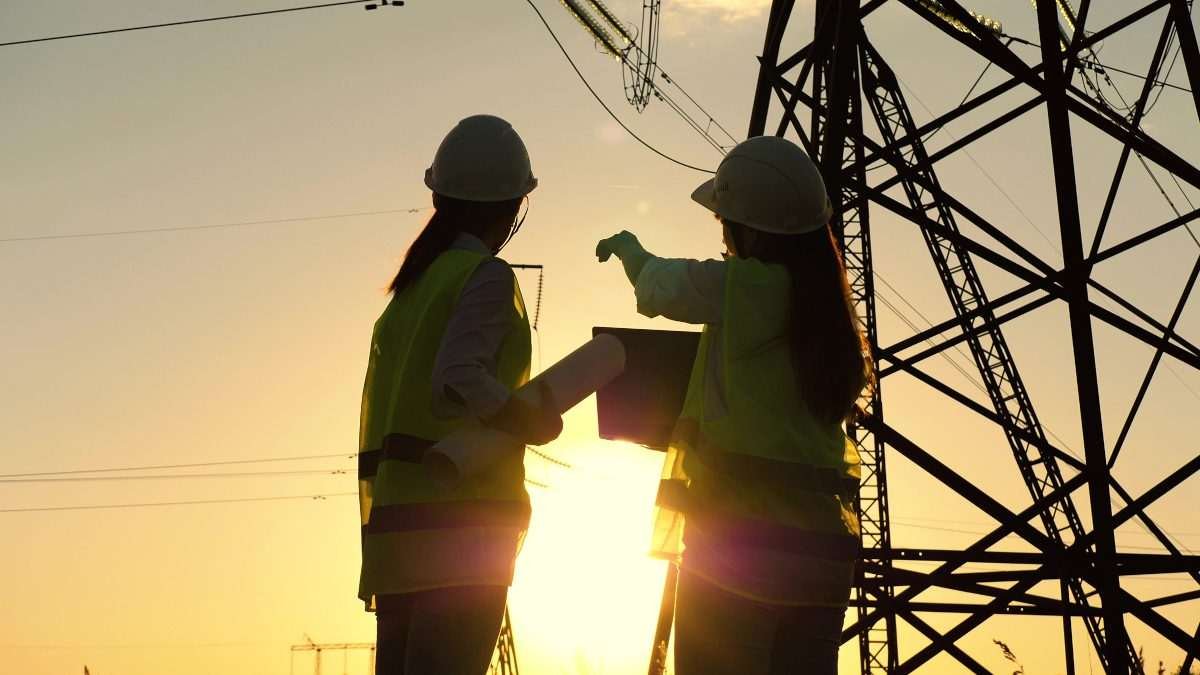 Two figures in hard hats silhouetted against bright dawn or dusk sky with electricity pylon tower visible in the background