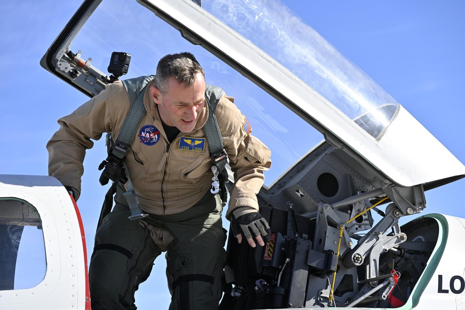 A test pilot, wearing a brown flight suit and gravity harness, stands under an open glass cockpit, looking over the side of a white aircraft as he prepares to exit via ladder.