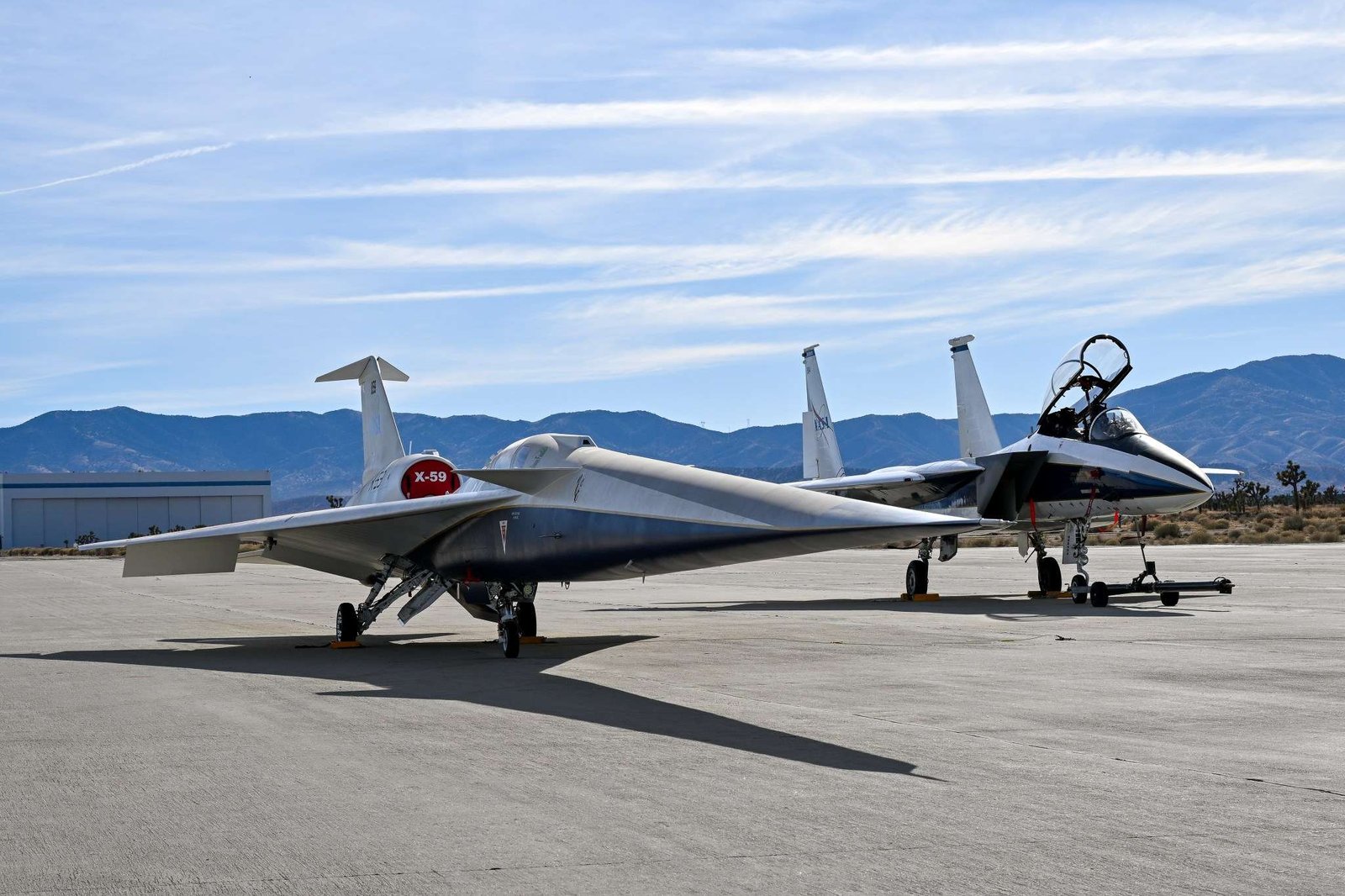 A NASA F-15 aircraft sits 20 feet off the left side of the X-59 aircraft, with a white hangar and hills in the background, during electromagnetic interference testing.