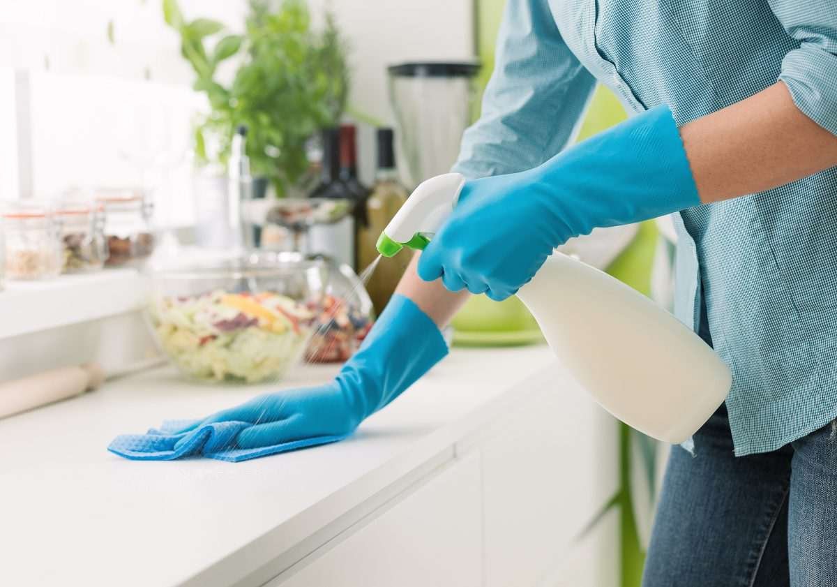 Rubber-gloved female hands using spray-based cleaner on a kitchen surface
