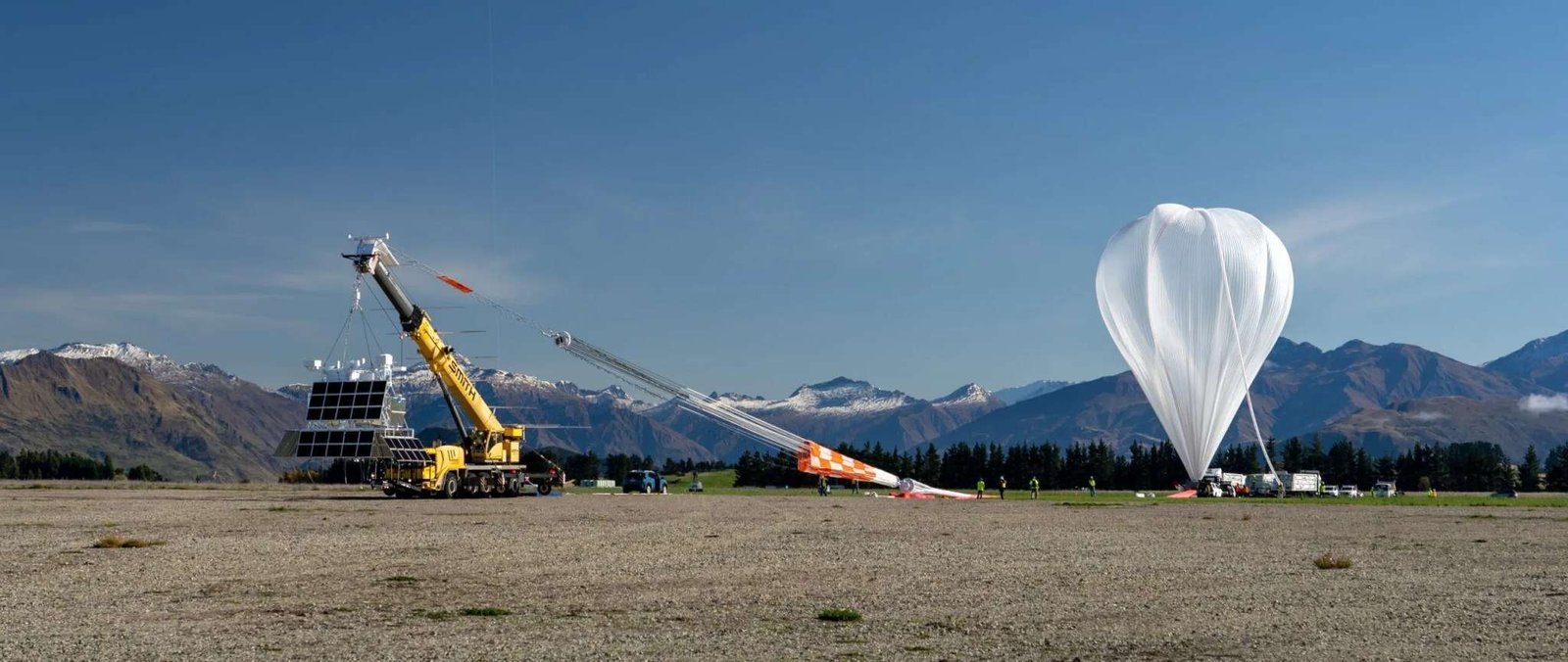 A large scientific balloon is tethered to the ground as it inflates before liftoff in New Zealand. The super pressure balloon is attached to an orange and white parachute, and a scientific payload is suspended from a mobile launch crane.