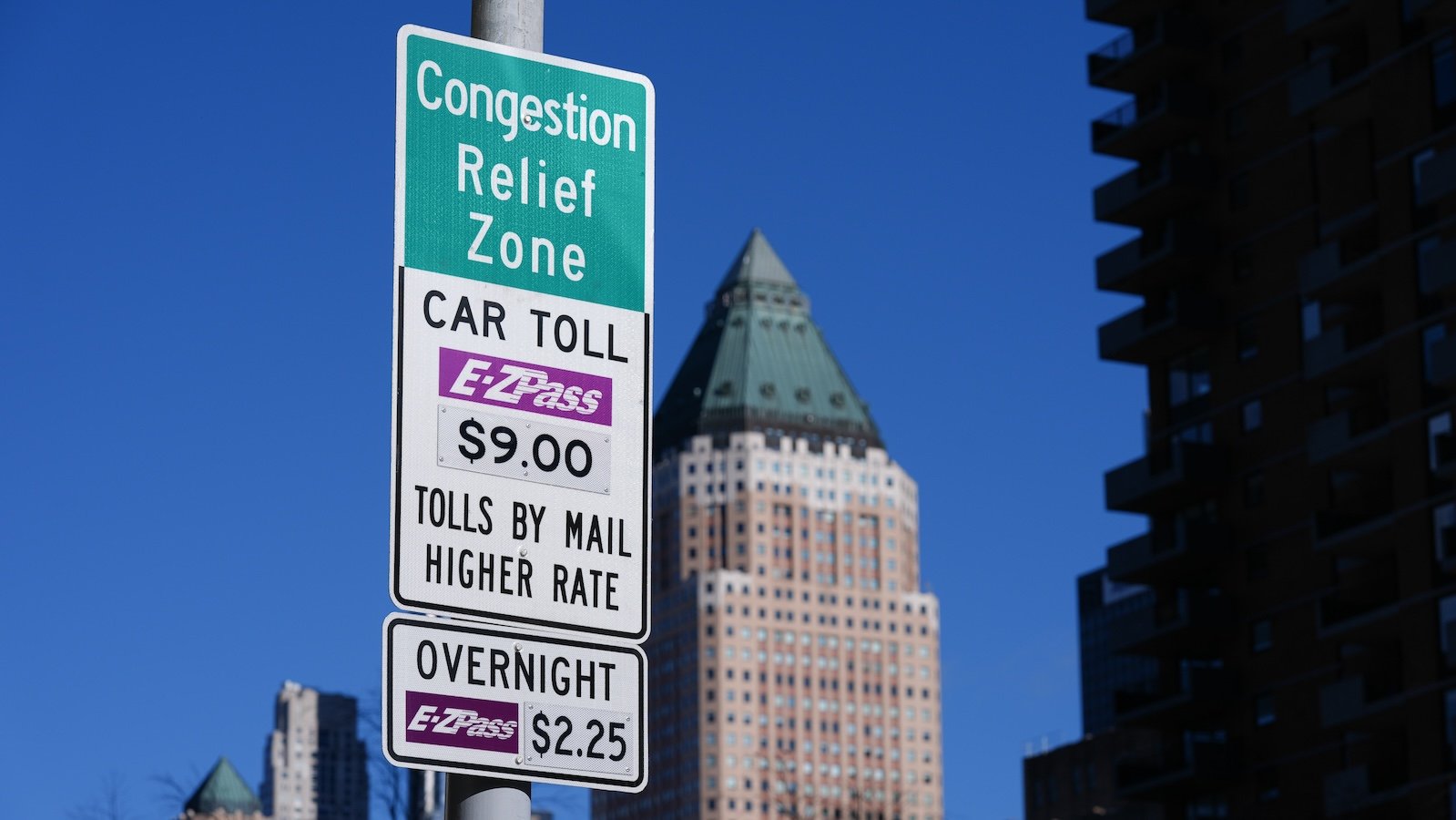 Close-up of a sign announcing congestion pricing tolls in New York City, with a blue sky and building in the background