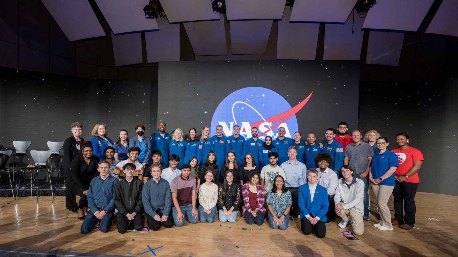 A group of people pose in front of a NASA meatball with a starry background. The people in the first two rows are kneeling down. Most of the people standing in the back row are wearing blue flight suits.