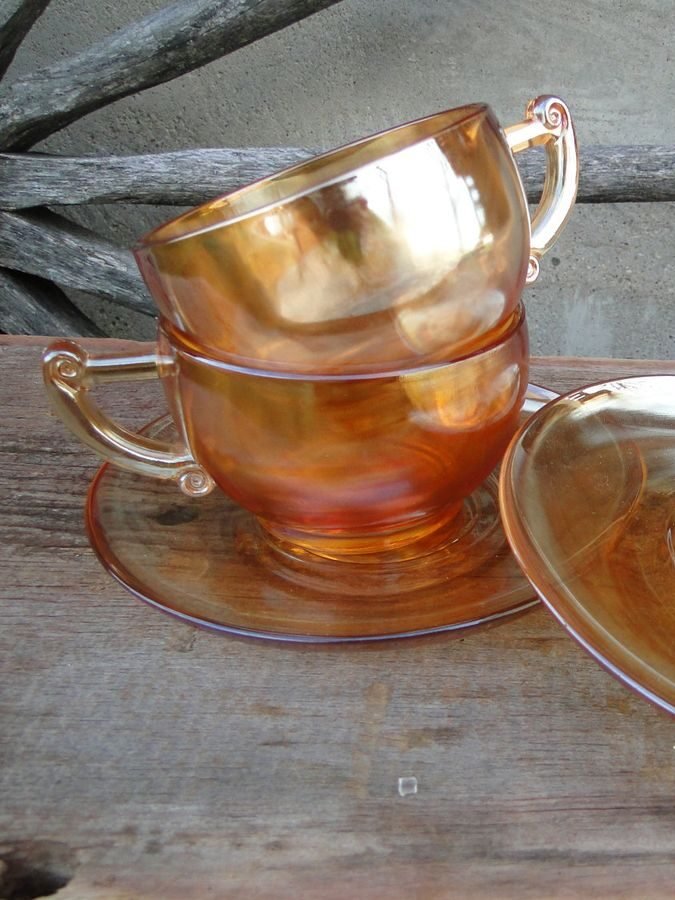 Two stacked amber glass teacups and saucers on a wooden table, with a rustic wheel and dried flowers in the background.