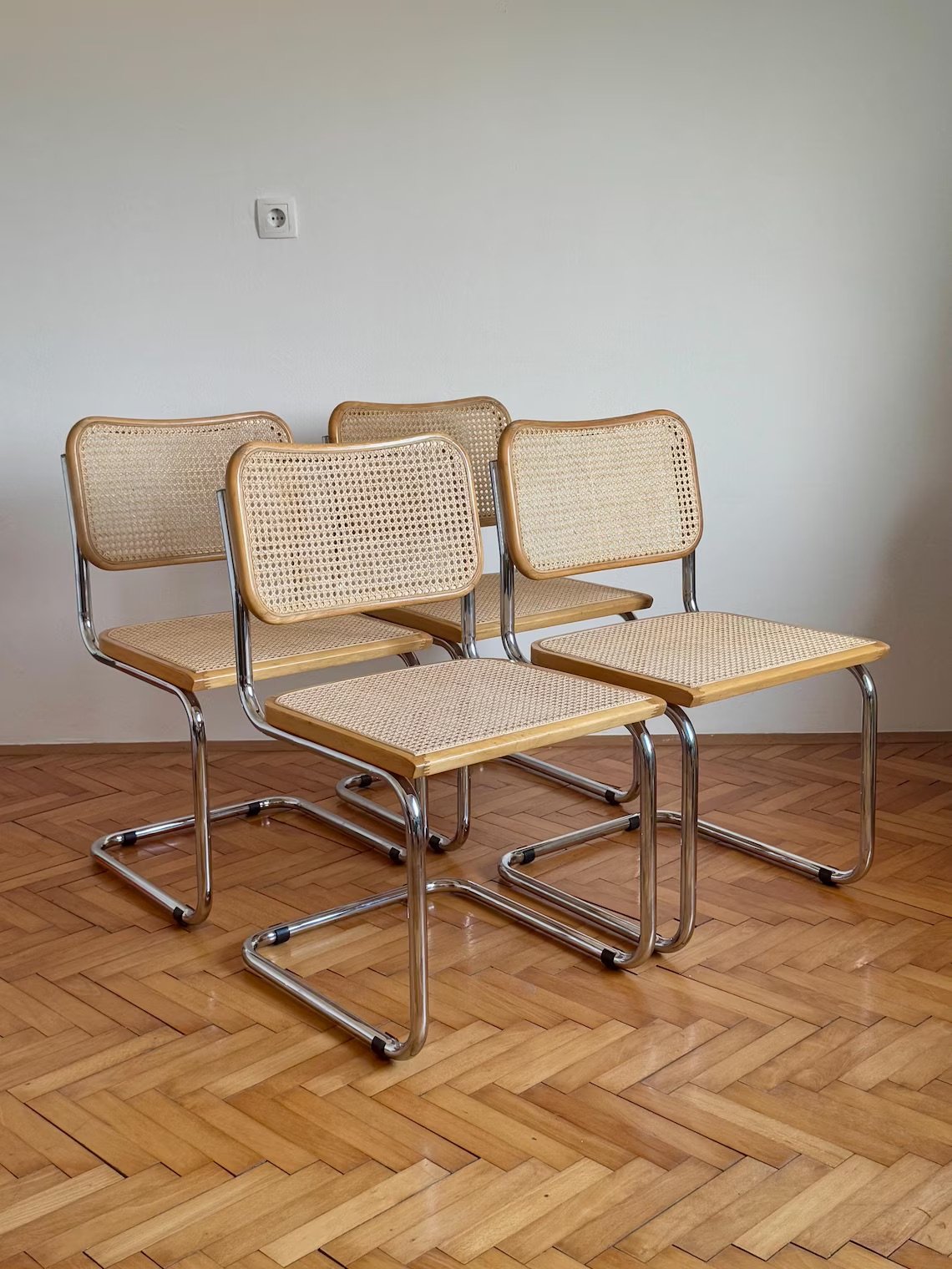 Four cane-backed chairs with chrome legs are arranged in pairs on a wooden parquet floor against a white wall.