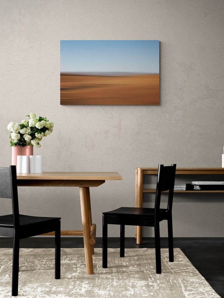 Minimalist dining area with a wooden table, two black chairs, a floral centerpiece, and a blurred landscape painting on the wall. Neutral colors dominate the decor.