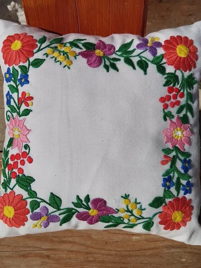 A white cushion with a floral embroidery pattern along the edges, featuring red, pink, yellow, and blue flowers, set on a wooden surface.