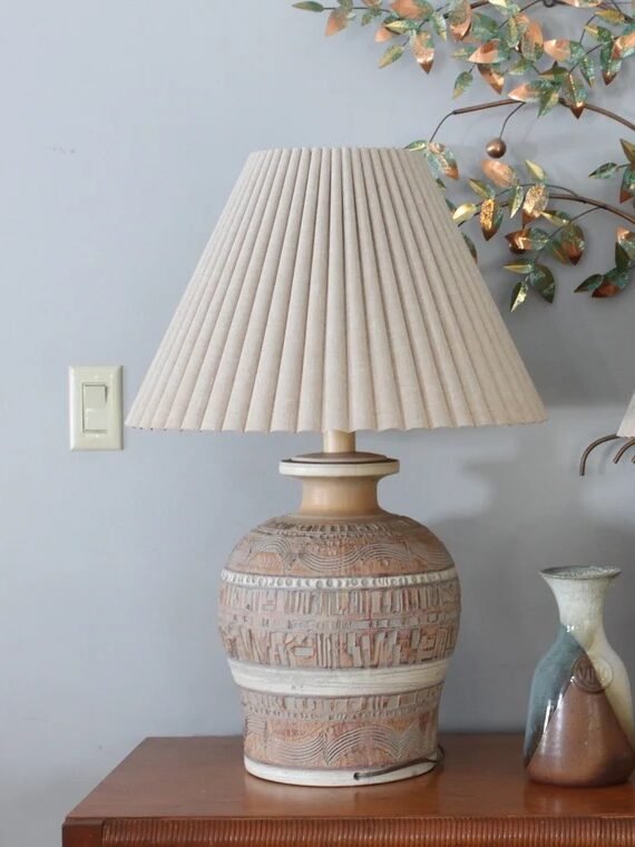 Two table lamps with pleated shades sit beside a small decorative vase on a wooden surface, with a metallic leaf wall decoration in the background.