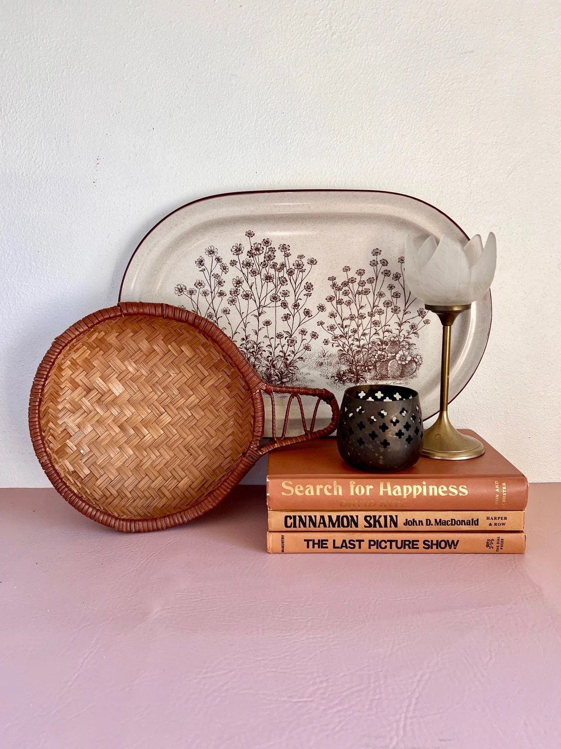 Stack of three books with a decorative plate and basket against a wall. A candle holder and a small lamp sit on top of the books.