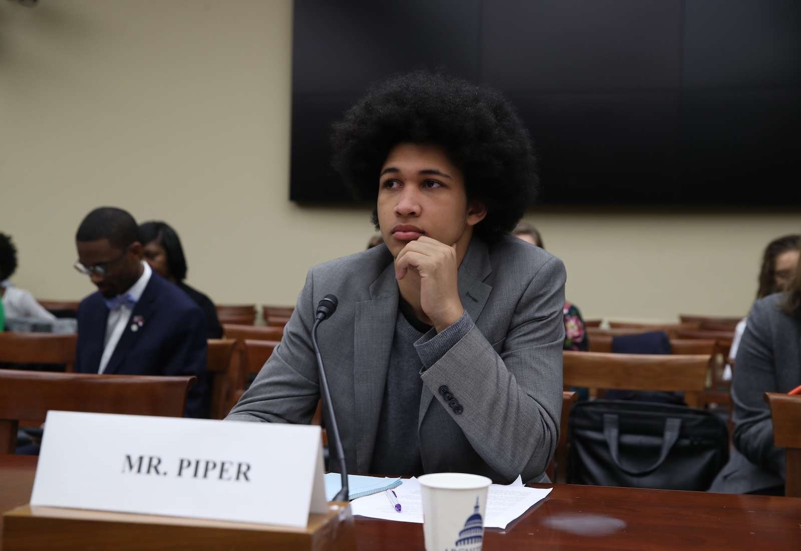 A young man wearing a suit sits at a table with a name tag reading "Mr. Piper" in front of him