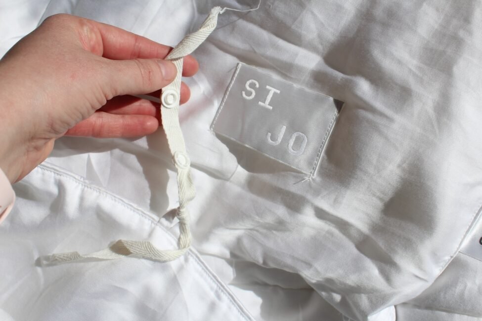 A hand holds a white tied ribbon on a white fabric, with a visible tag that says "SI JO.