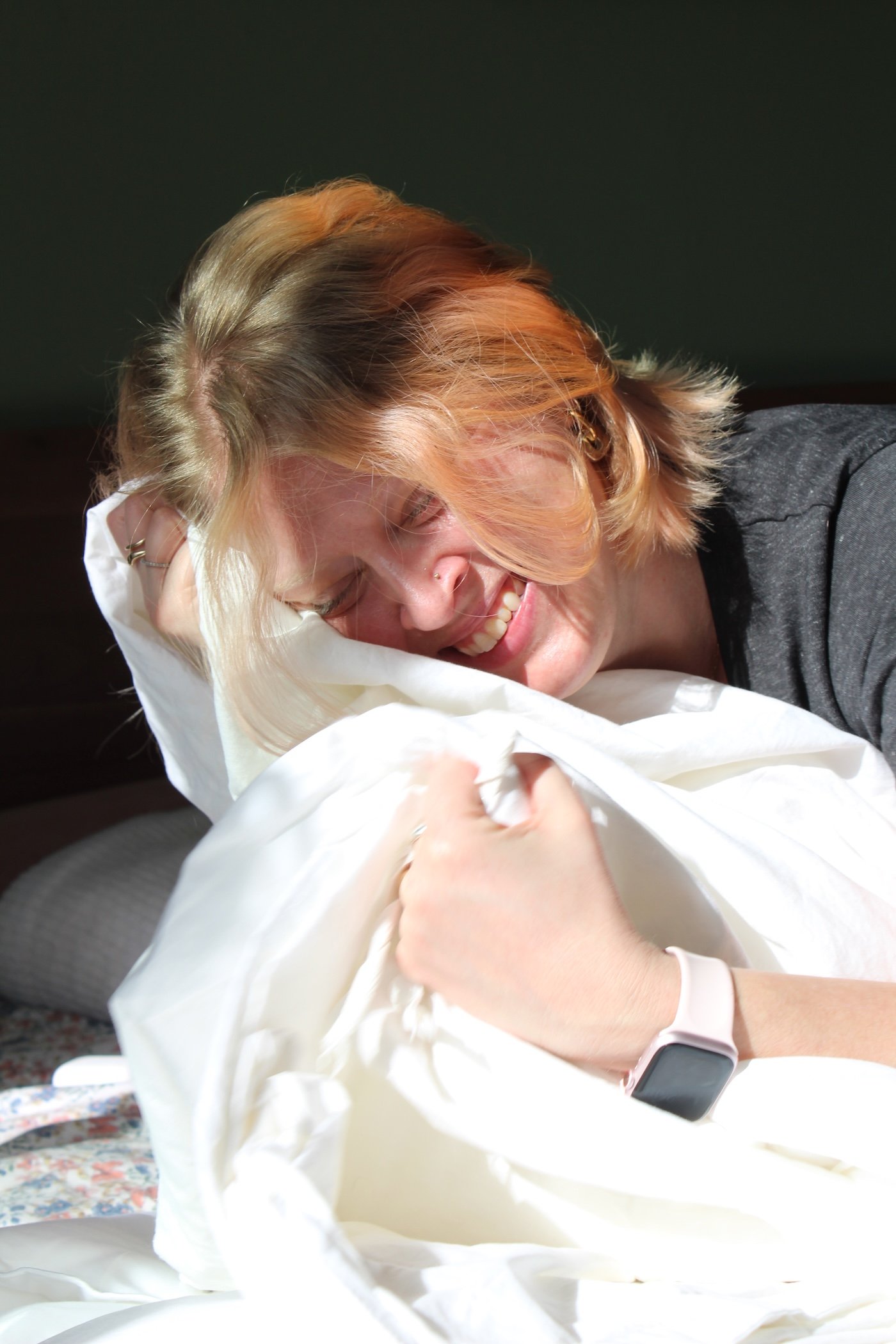 A person with shoulder-length hair is lying on a bed, smiling and holding a white pillow, in bright sunlight. They are wearing a dark shirt and a smartwatch.