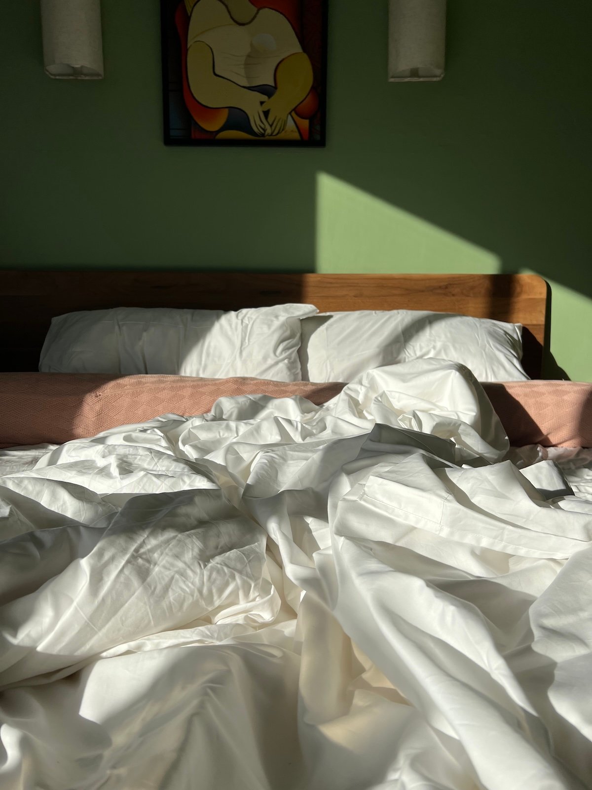 Unmade bed with white sheets in sunlight, underneath a painting of an abstract figure.