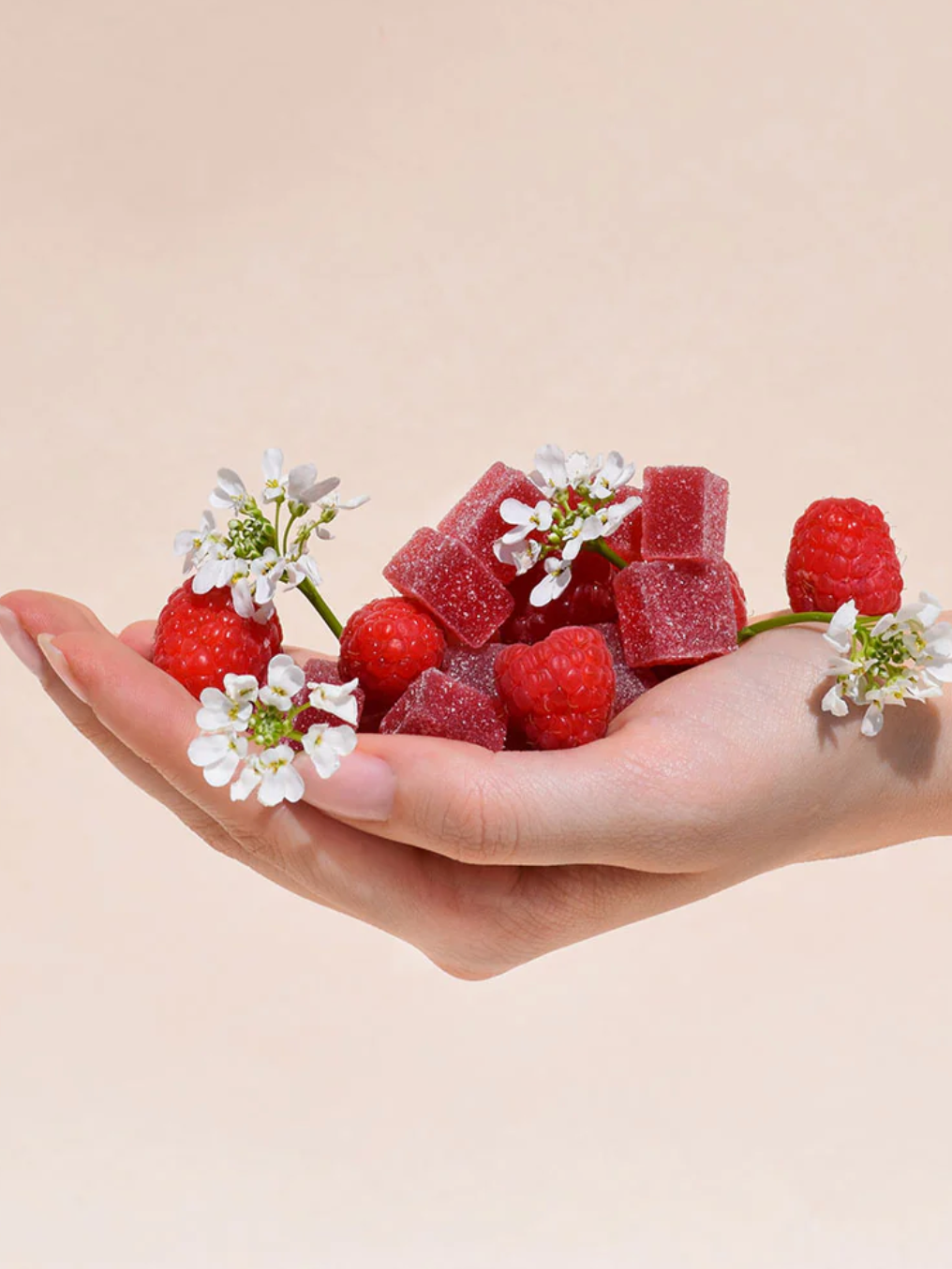 A hand holds red gummy cubes, fresh raspberries, and small white flowers against a plain, light beige background.