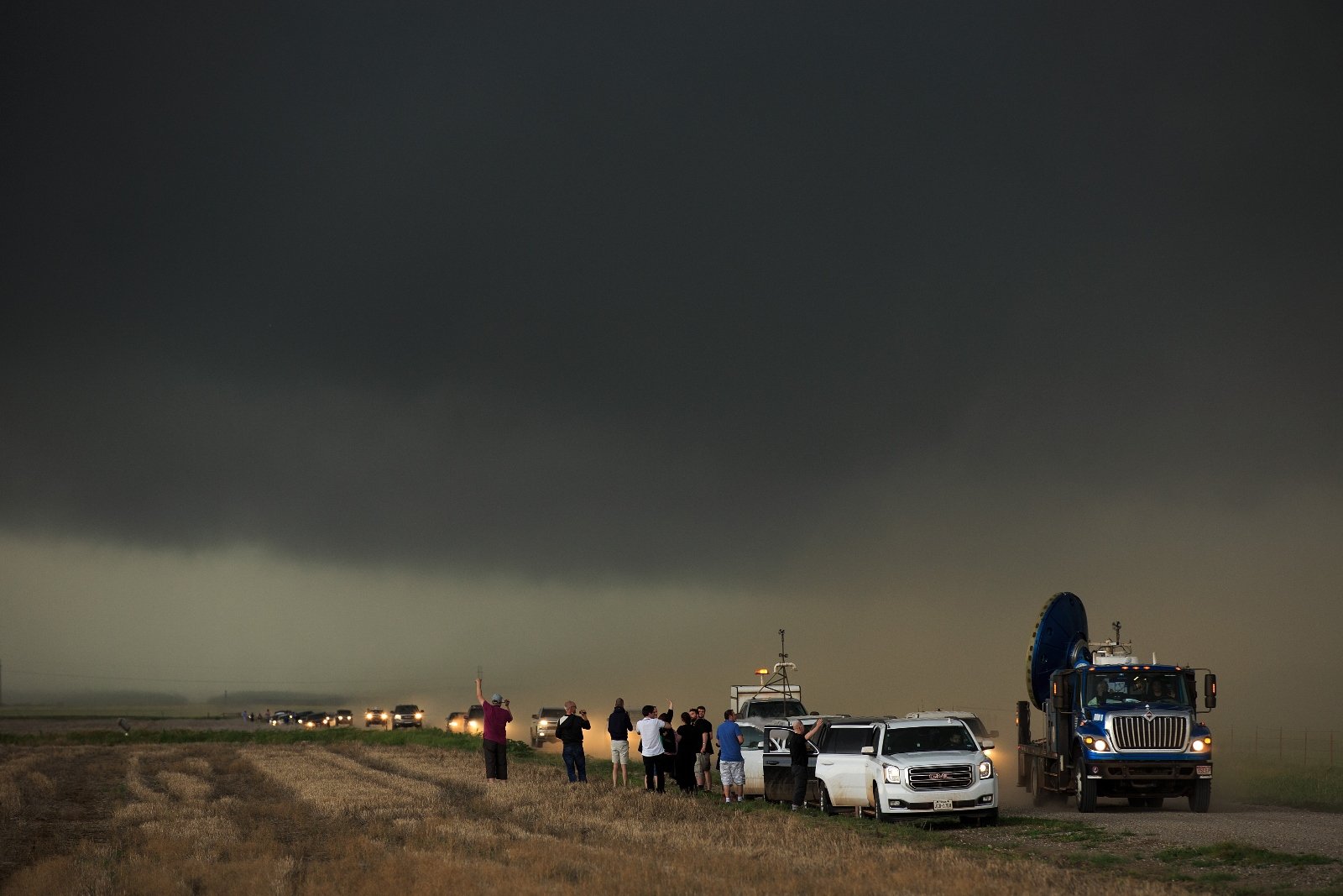 A caravan of cars and trucks on the side of the road, as people stand outside and look up at a sky dark with storms.